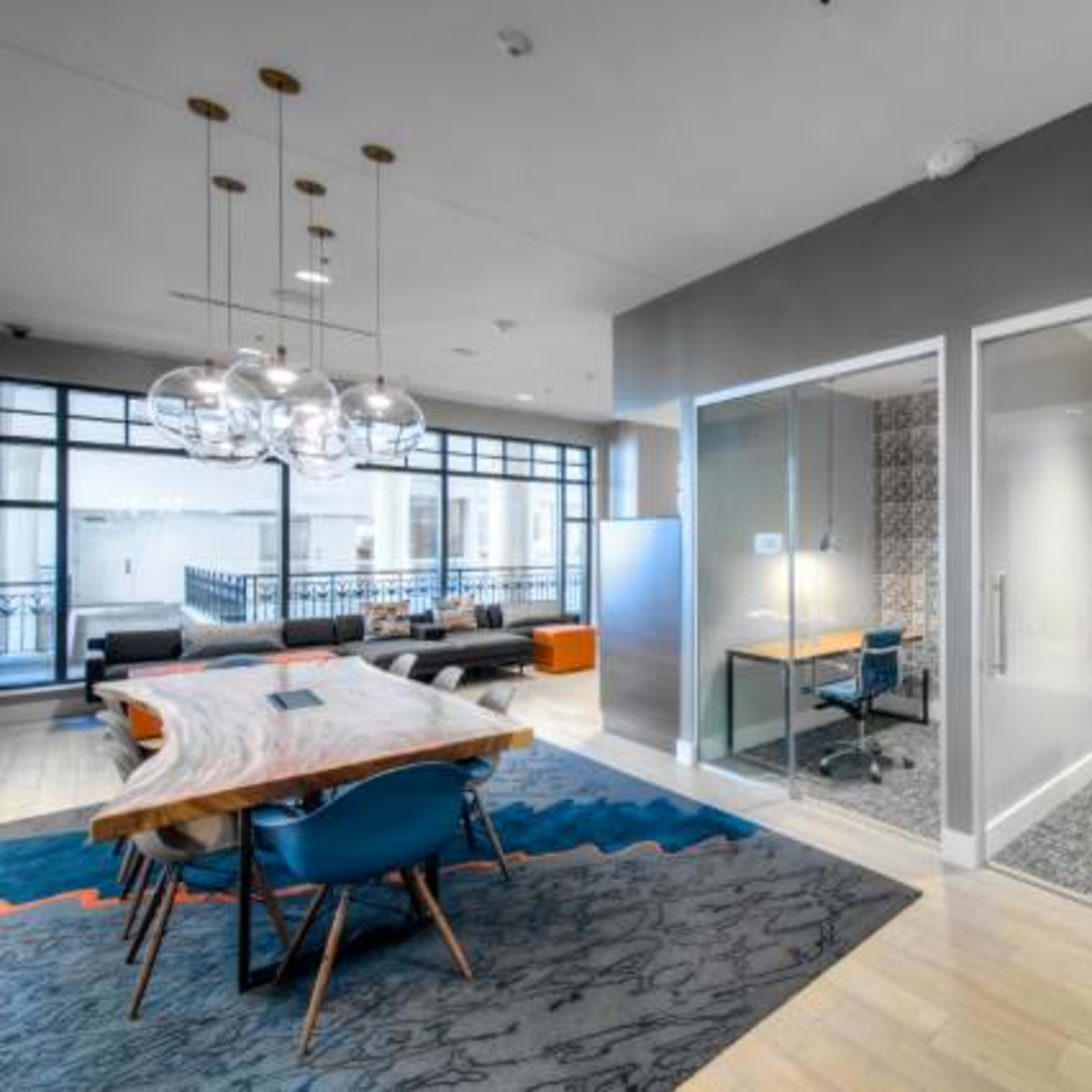 A modern office space featuring a large wooden table with blue chairs, a patterned area rug, and two glass-enclosed workstations.
