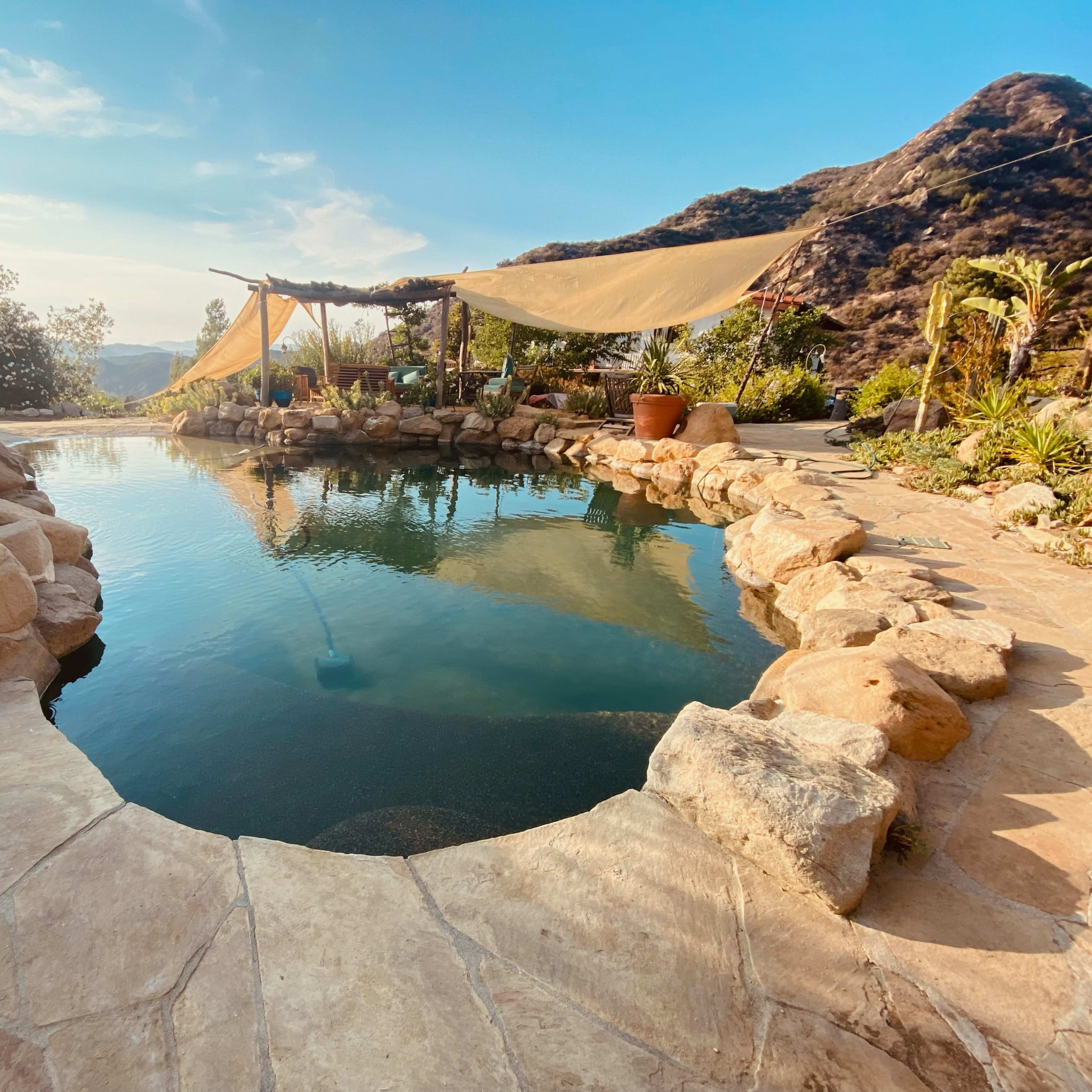 A natural stone pool surrounded by lush vegetation and a shaded seating area, set against a backdrop of rolling hills.