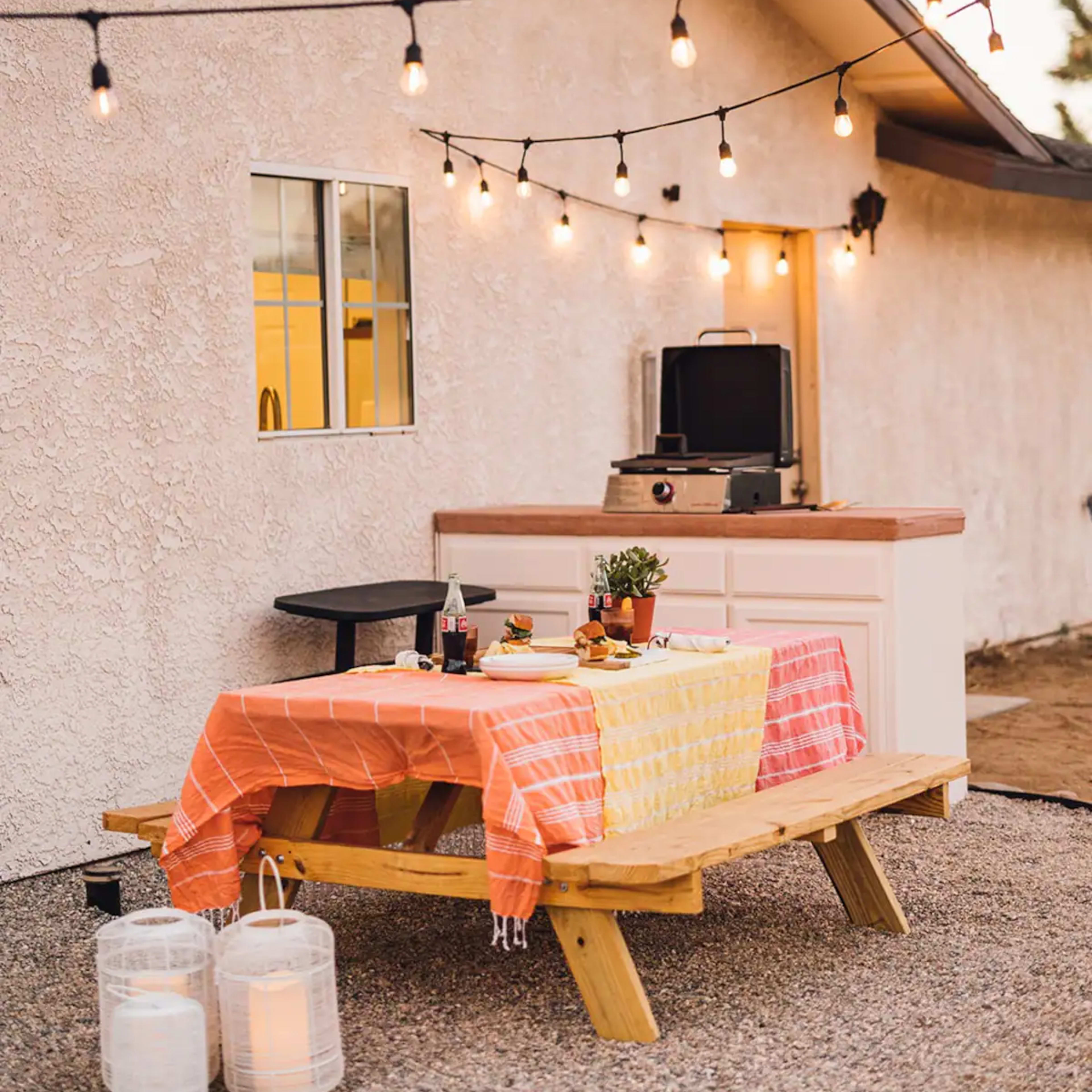 A backyard setting features a wooden picnic table with colorful tablecloths, a grill on a countertop, and string lights overhead.