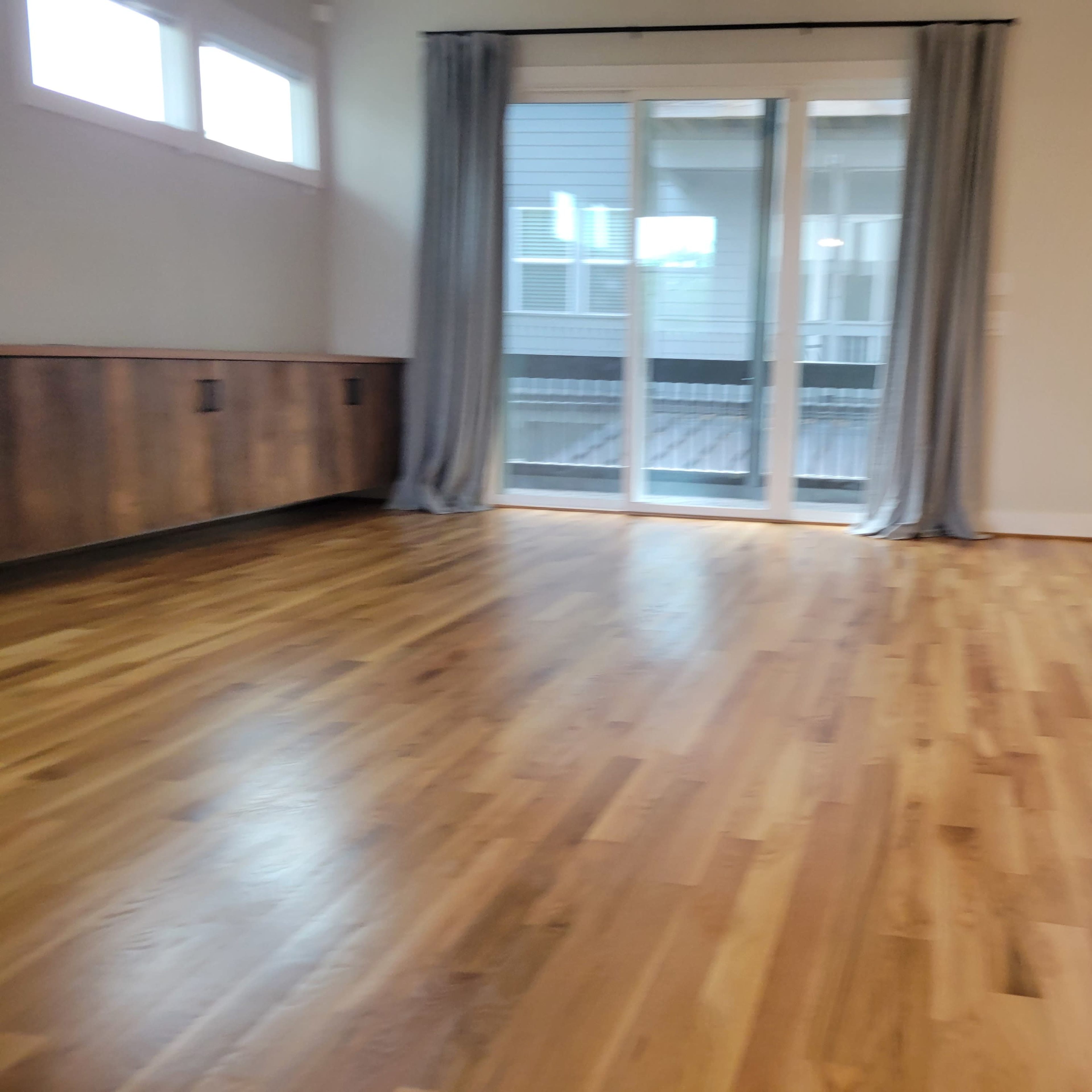 A spacious room with wooden floors, large sliding doors that lead to a balcony, and gray curtains on either side.