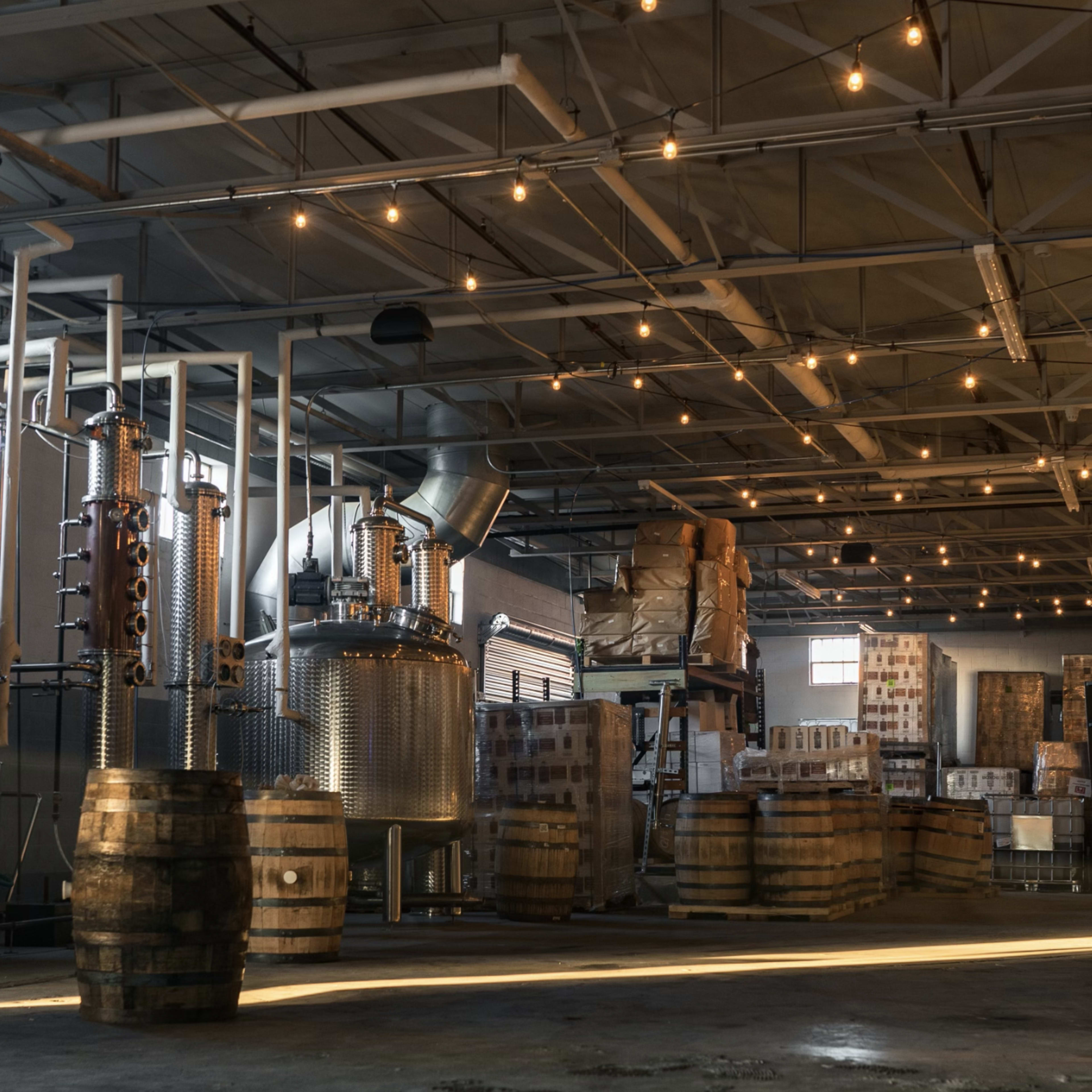 A distillery interior featuring copper and stainless steel stills, wooden barrels, and stacked boxes under rows of overhead lights.