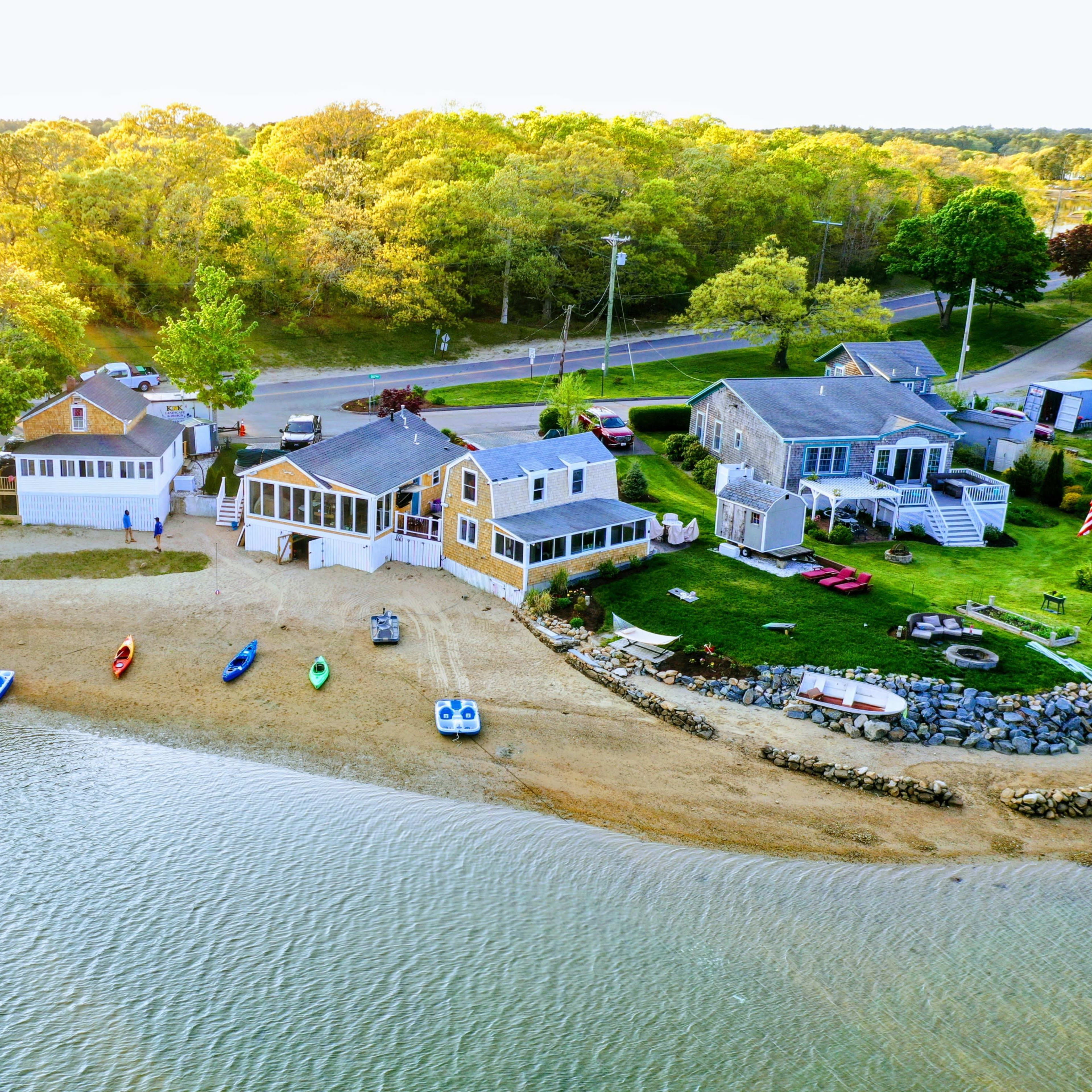 A coastal area with several houses alongside a sandy beach, where colorful kayaks and paddle boats are lined up by the water's edge.