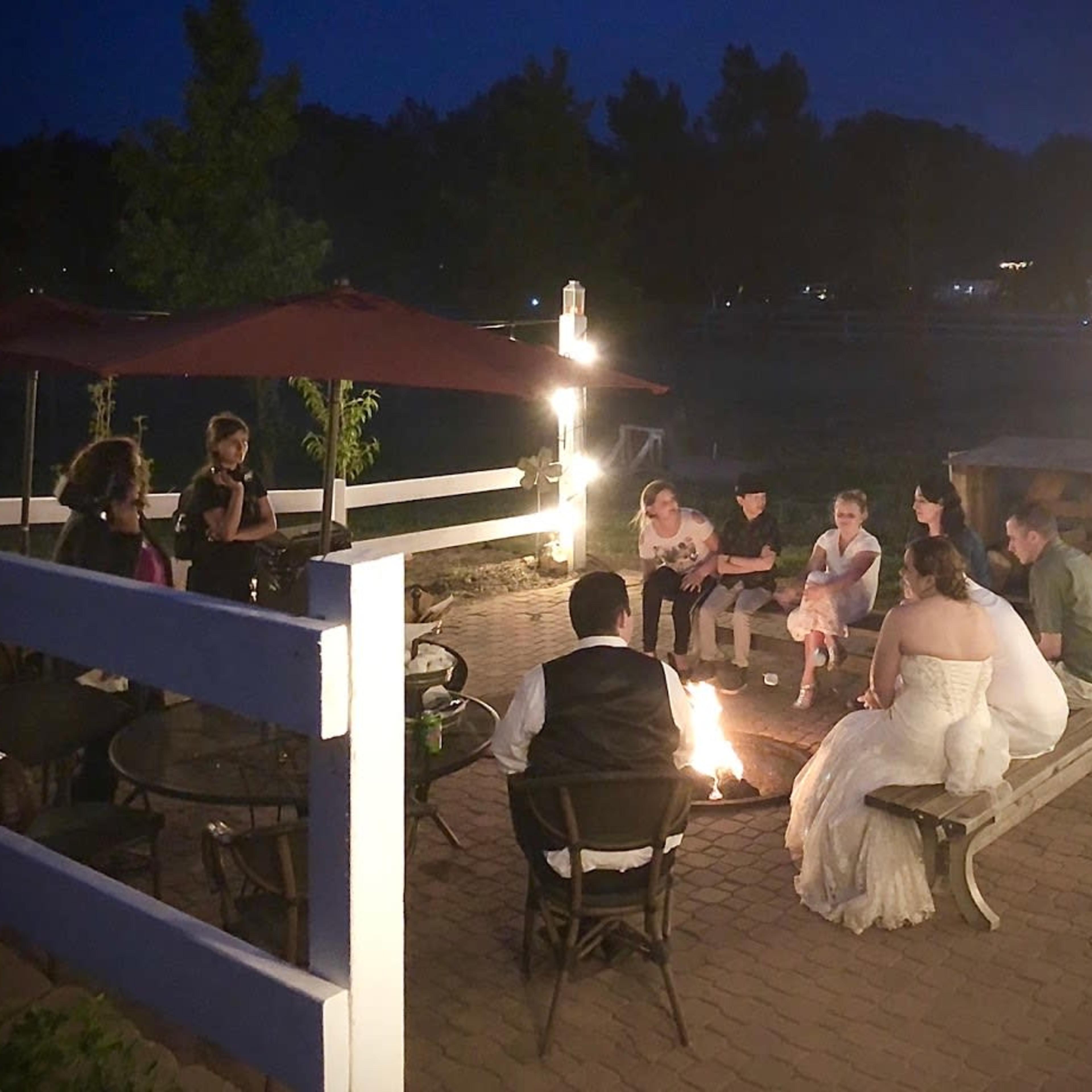 A group of people gather around a fire pit at night, seated on benches and chairs near an outdoor patio.