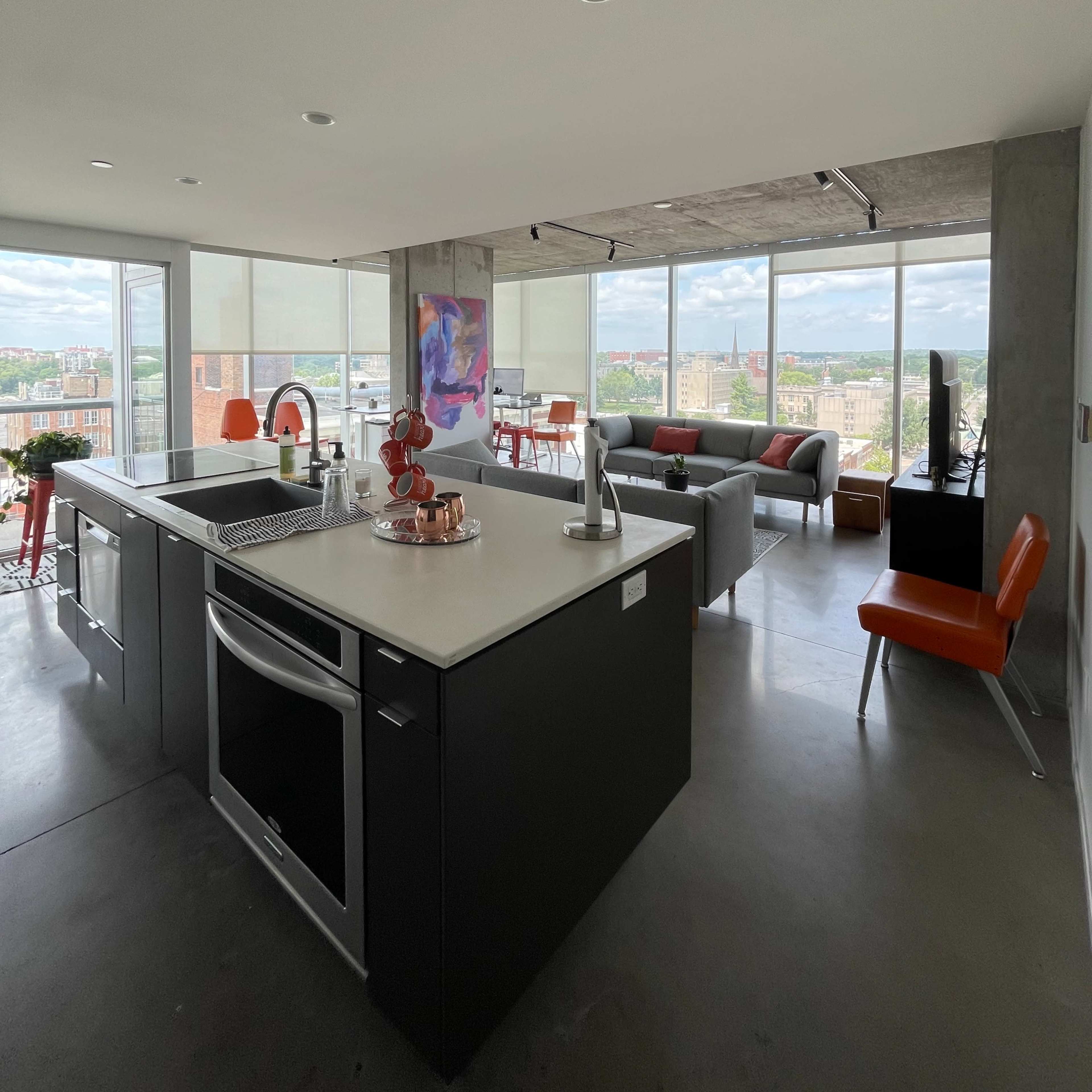 A modern, open-concept apartment features a kitchen with a large island and floor-to-ceiling windows that offer a view of the city.