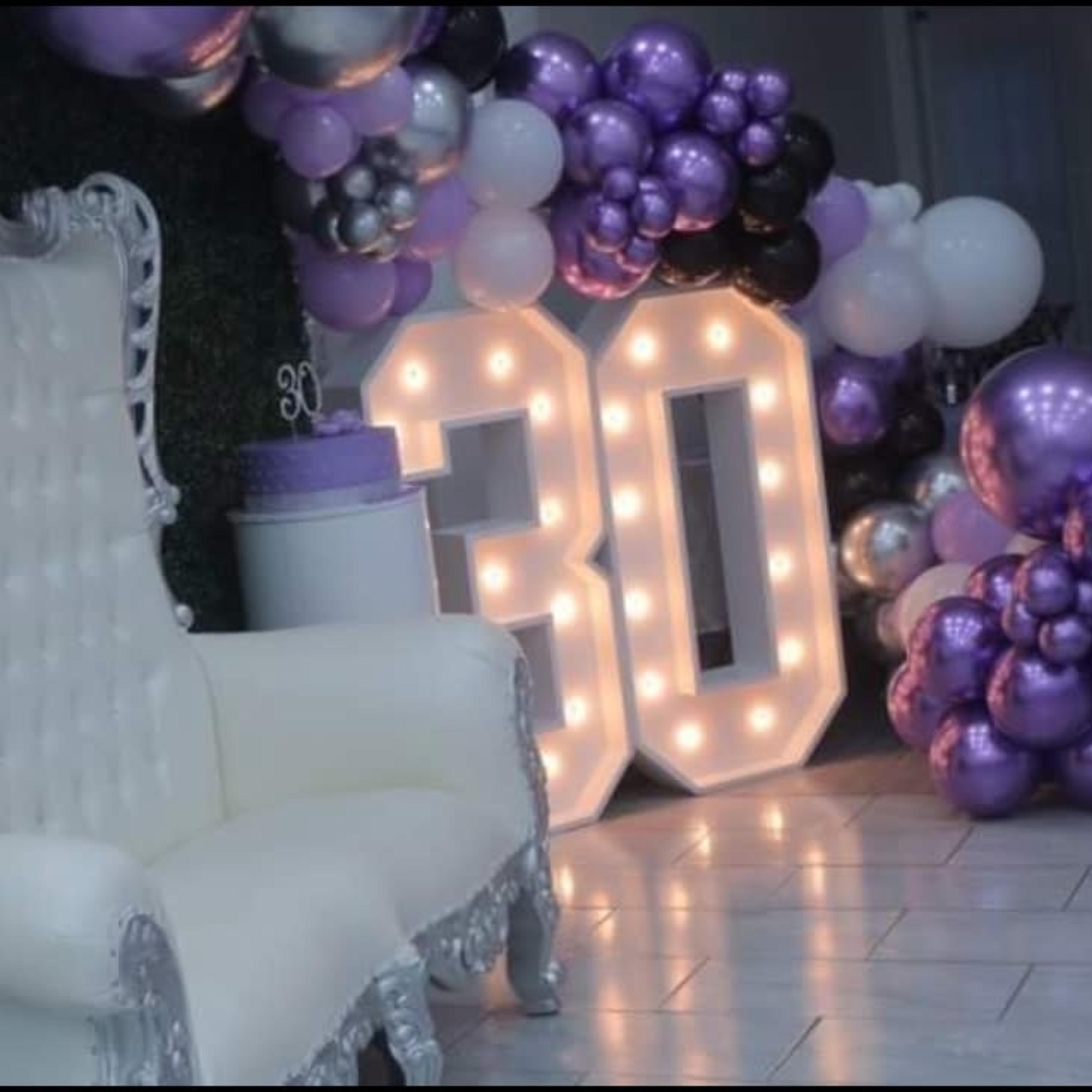 A large illuminated "30" display is surrounded by a cluster of purple, black, and white balloons, with an elegant white lounge chair positioned nearby.
