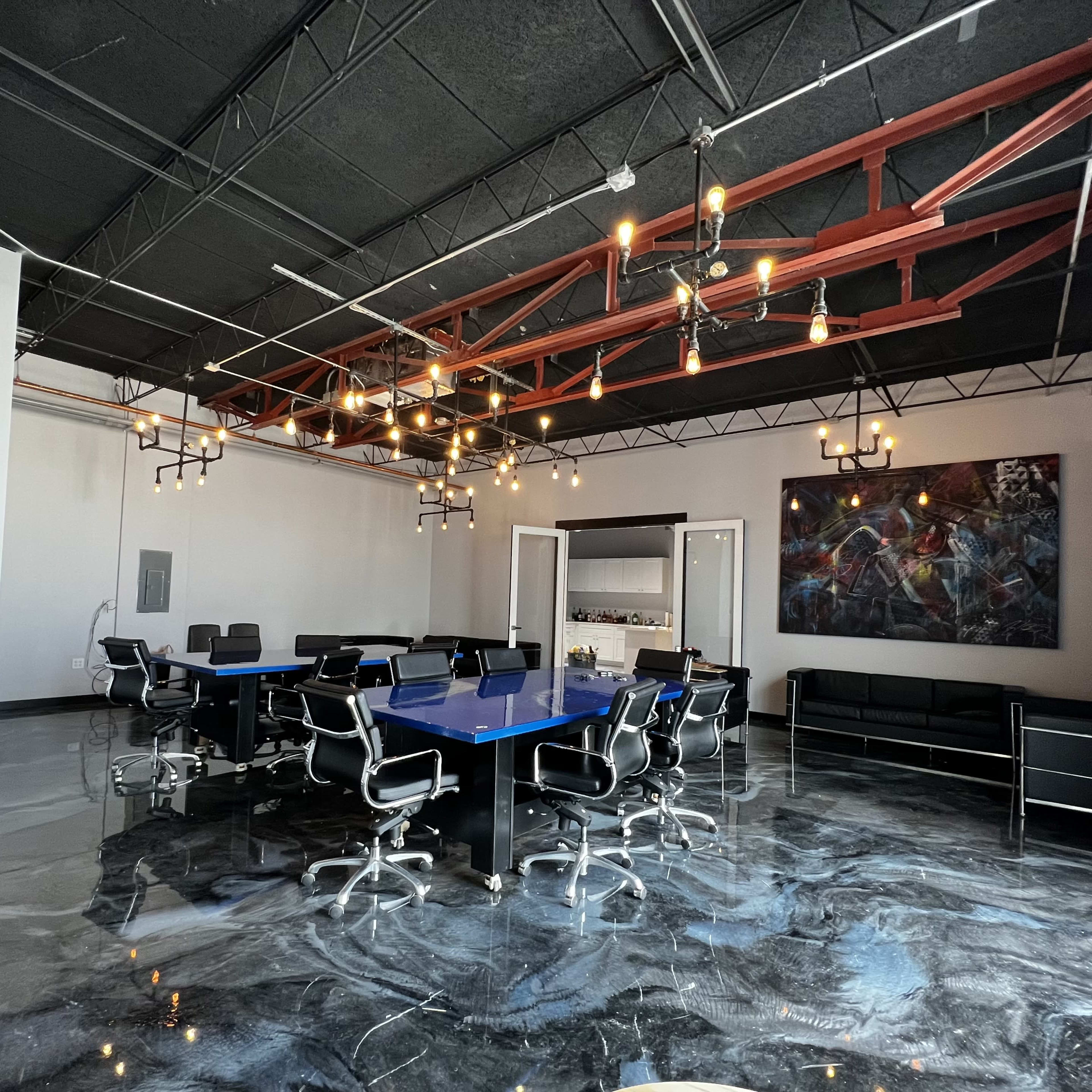 A modern conference room with a blue glass table, black swivel chairs, and a large abstract painting on the wall, illuminated by pendant lights.