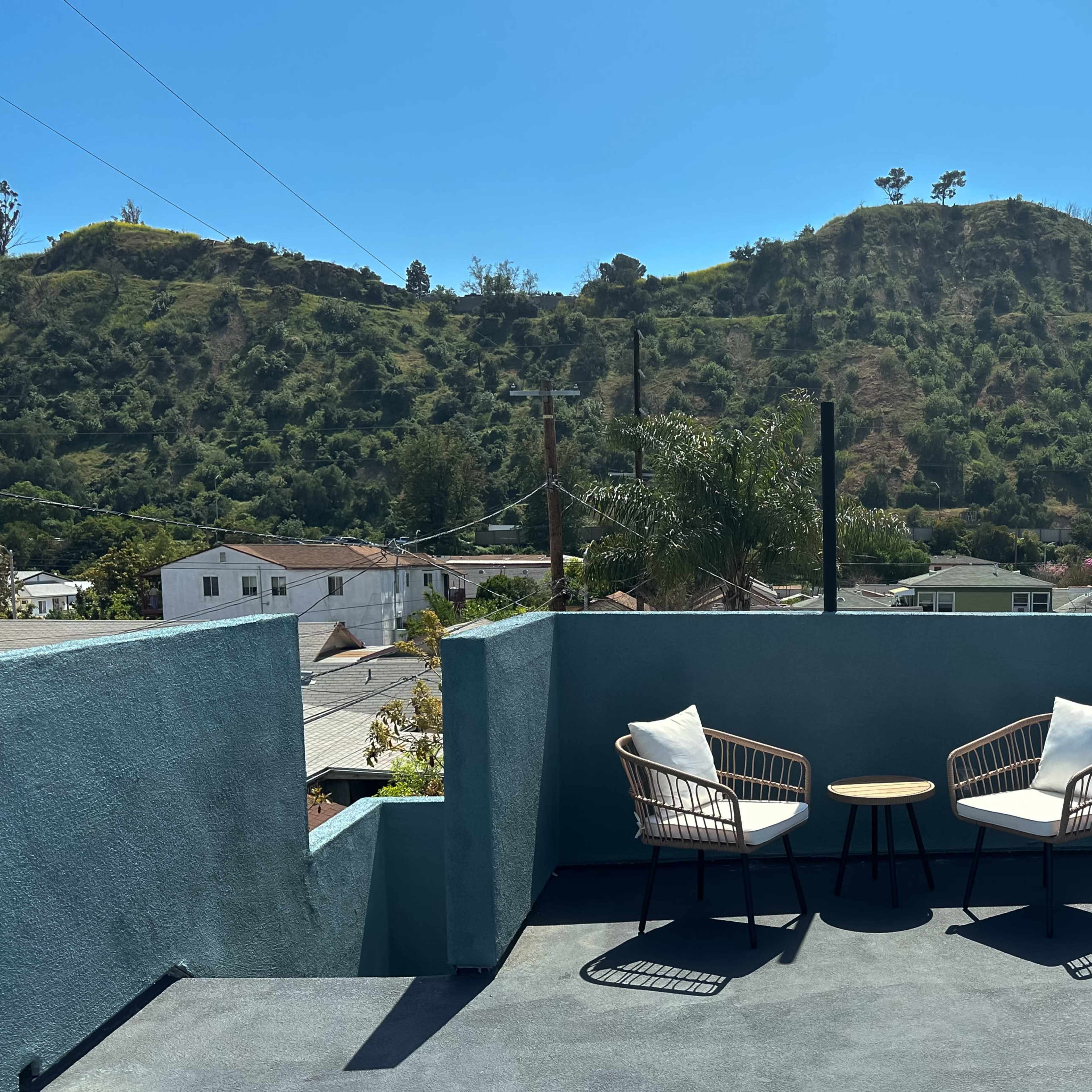 A rooftop terrace features two chairs and a small table, with a hillside and houses visible in the background.