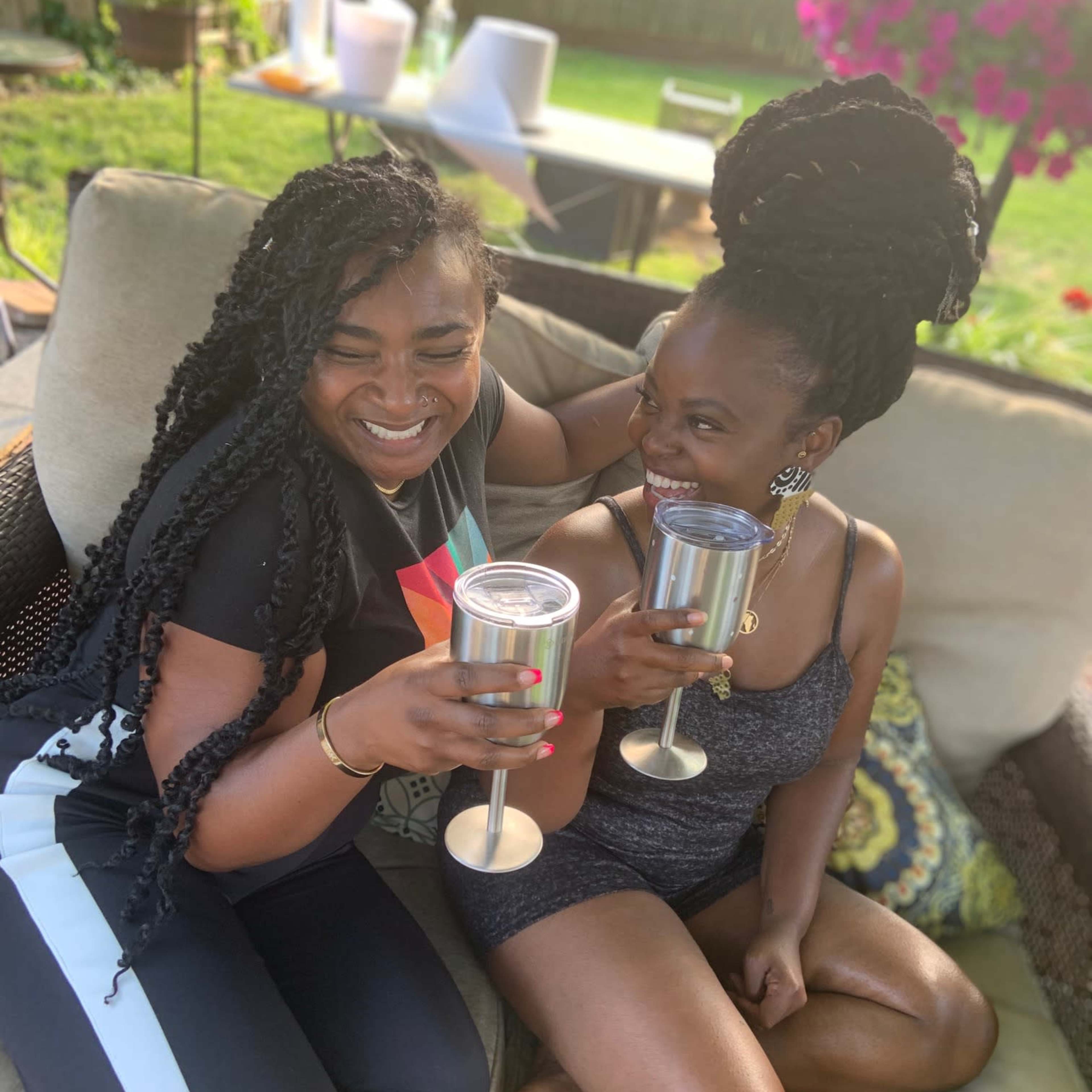 Two women are sitting on a patio couch, clinking stainless steel cups in celebration.
