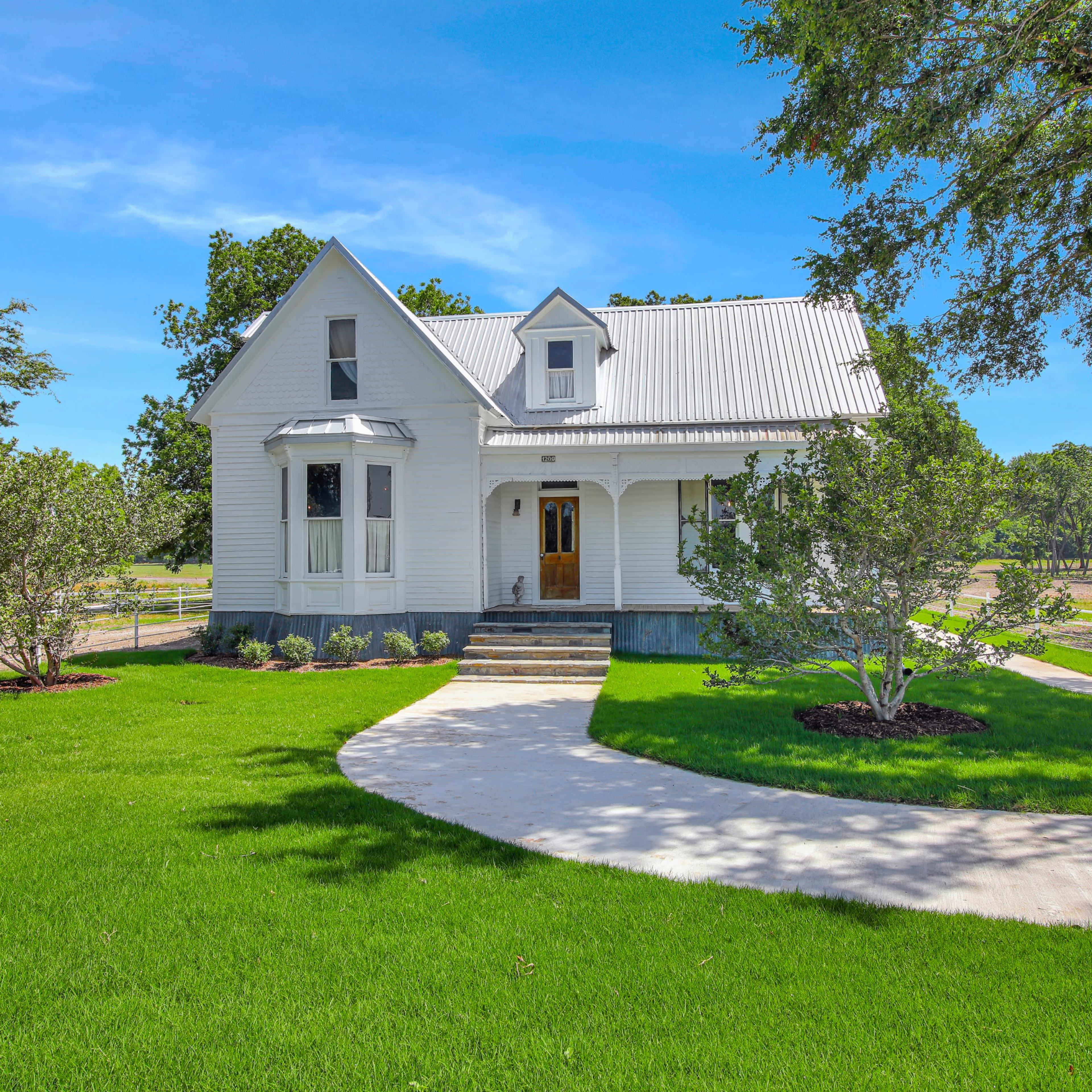 A white, two-story house with a metal roof surrounded by green grass and landscaped areas.