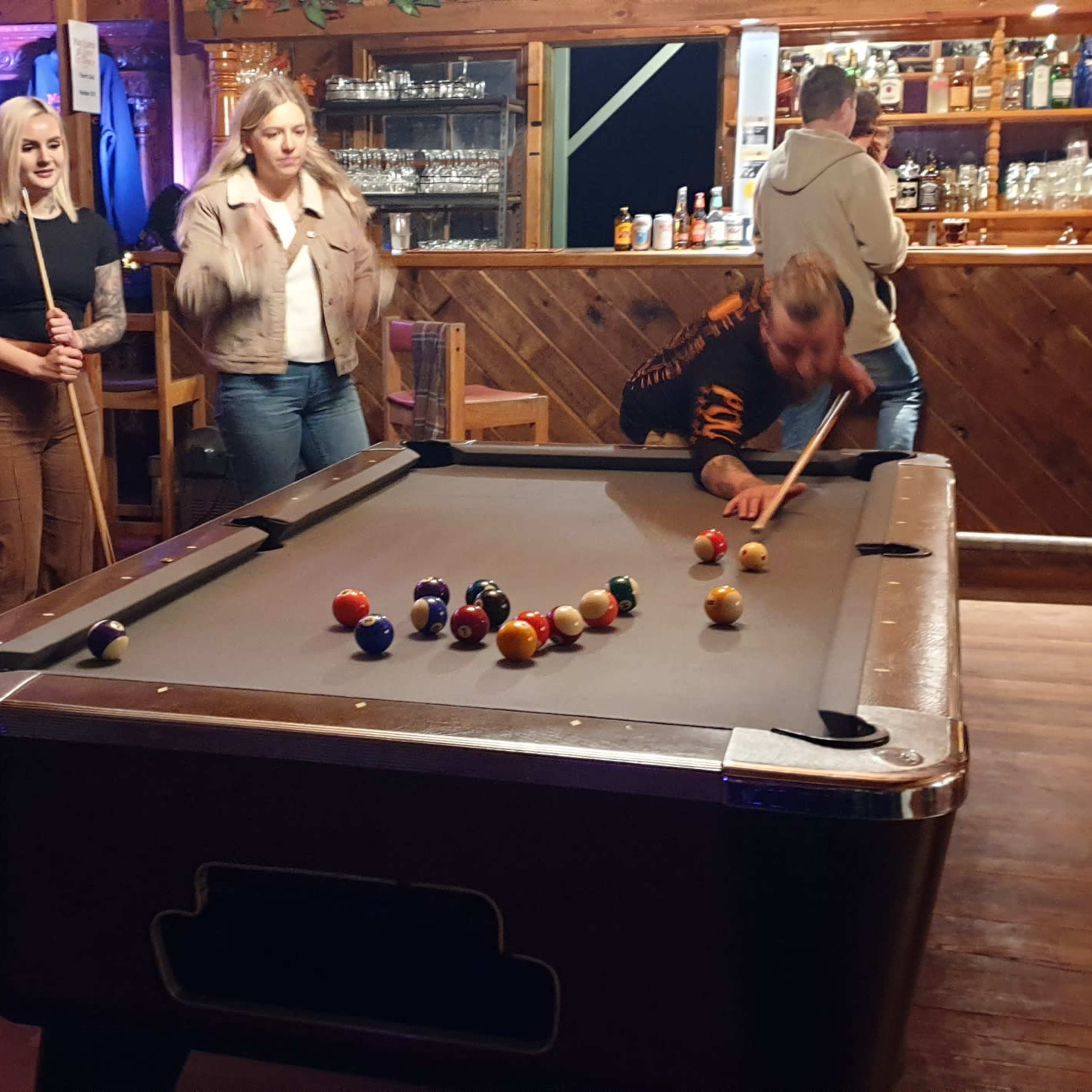 A group of people socialize in a bar while one man plays pool on a table.
