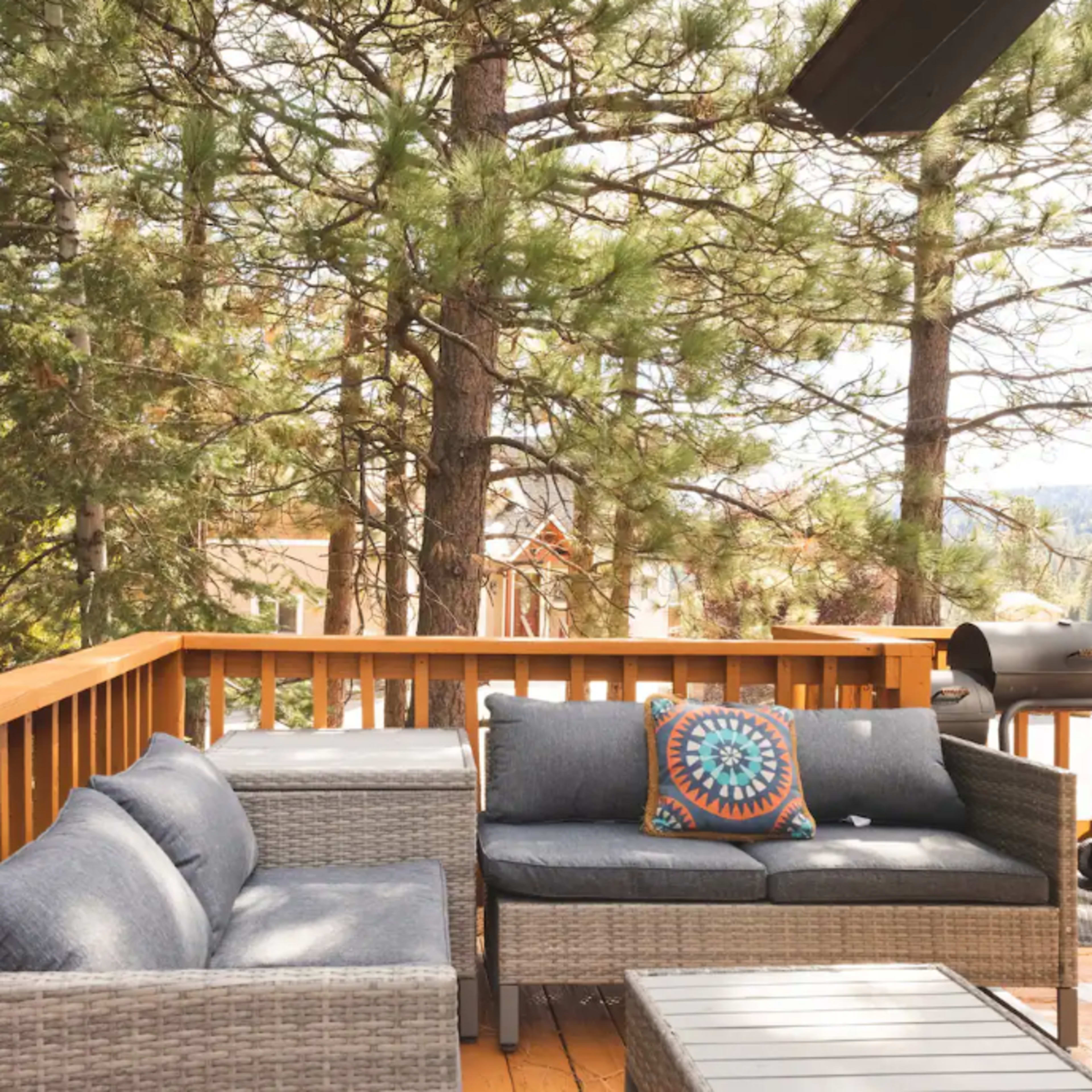 A deck area features wicker furniture, a decorative pillow, and a grill, surrounded by tall pine trees.