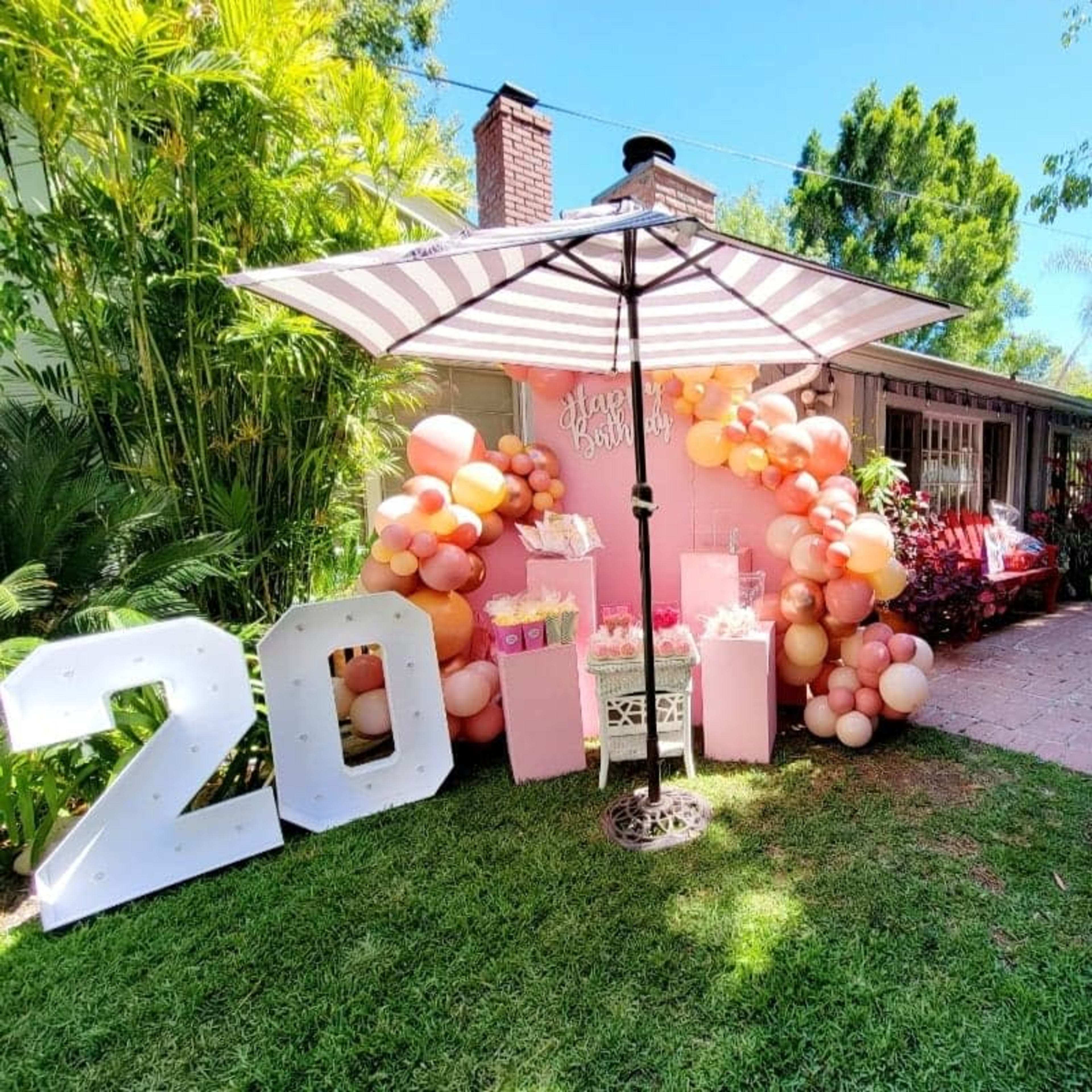 A birthday celebration scene features a large "20" display next to a festive balloon arrangement and gifts under a striped umbrella, with guests mingling near a pool in the background.