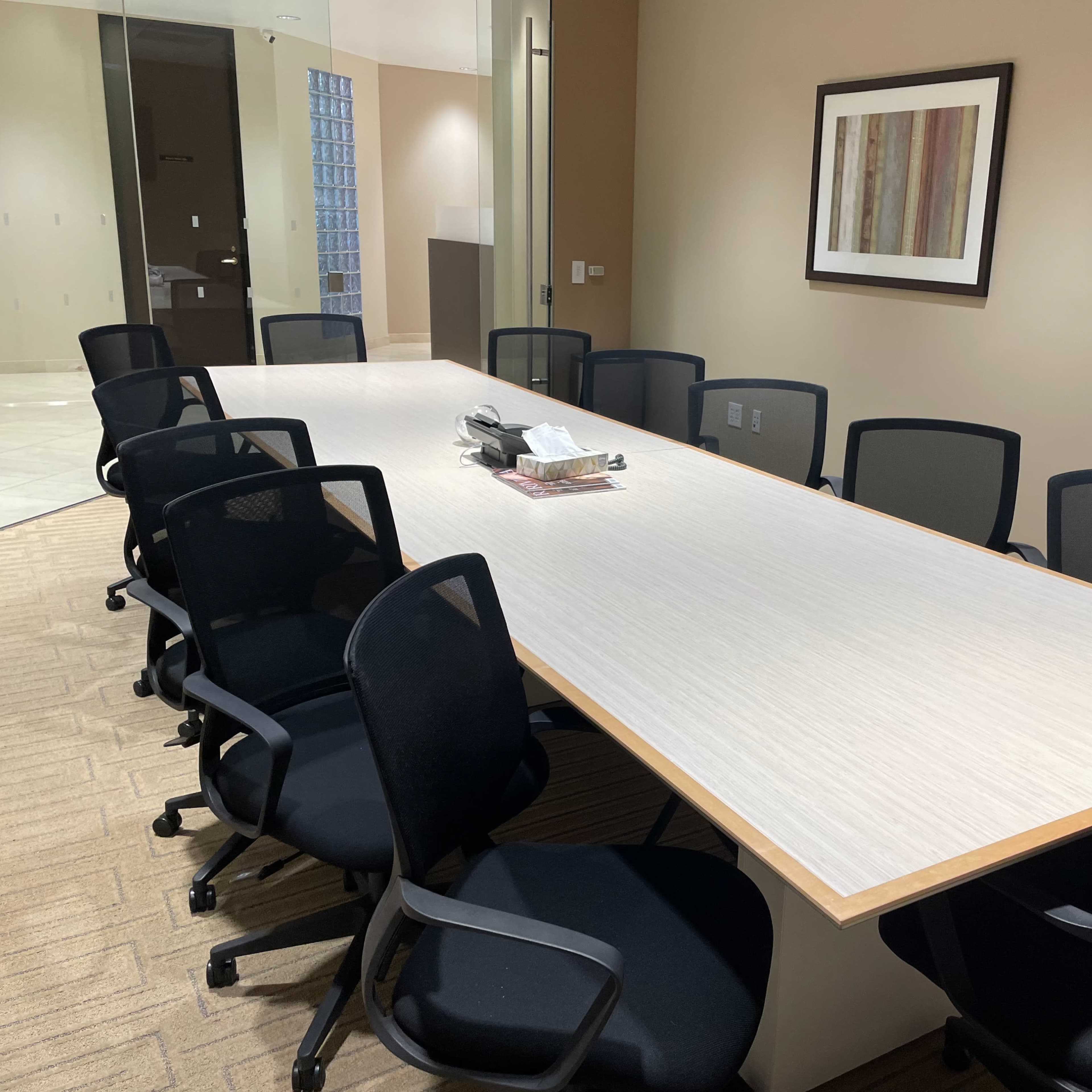 A long conference table is surrounded by black office chairs in a well-lit meeting room, flanked by two framed artworks on the walls.