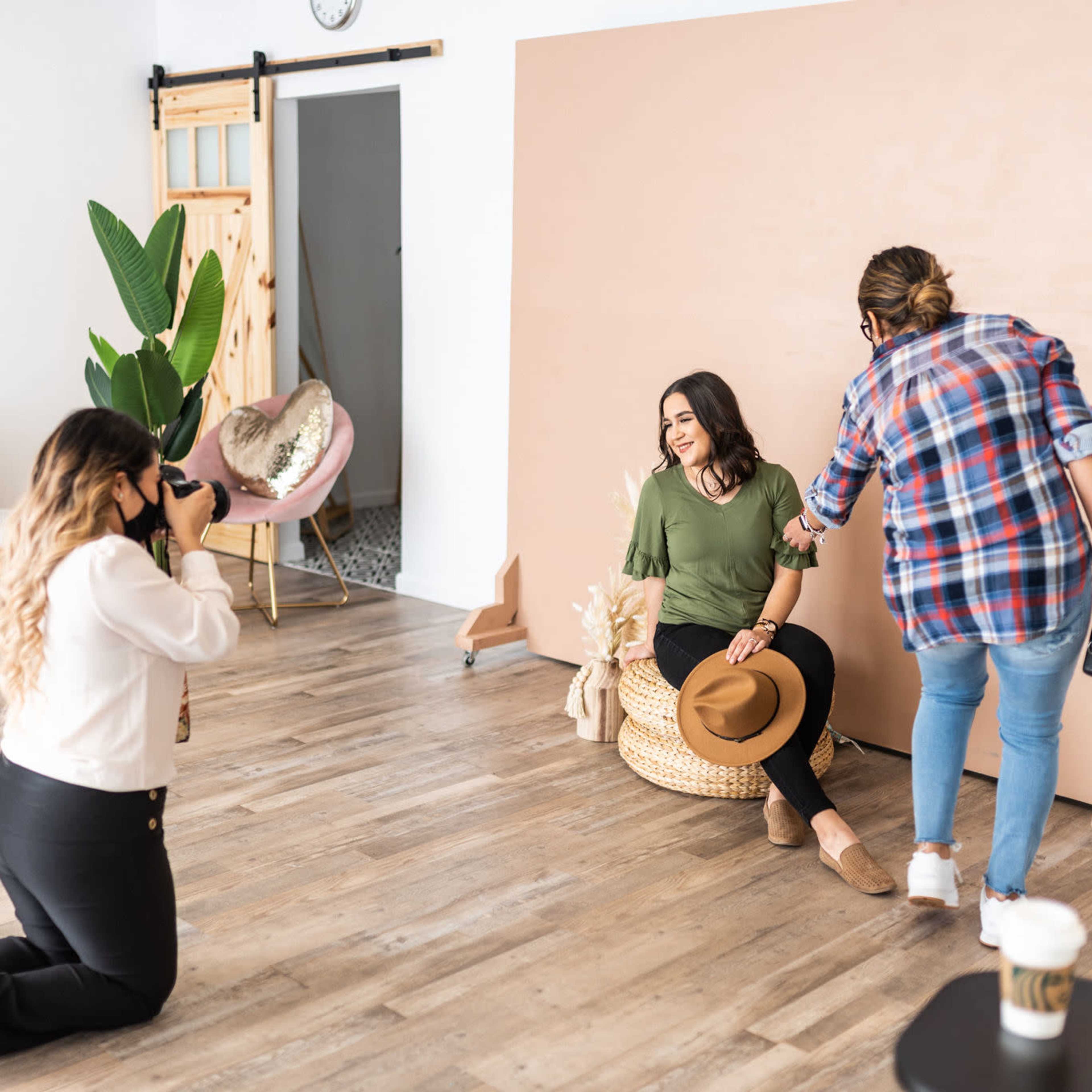 A photoshoot takes place in a studio with a model sitting on a chair, while a photographer captures the moment and a stylist adjusts the scene.