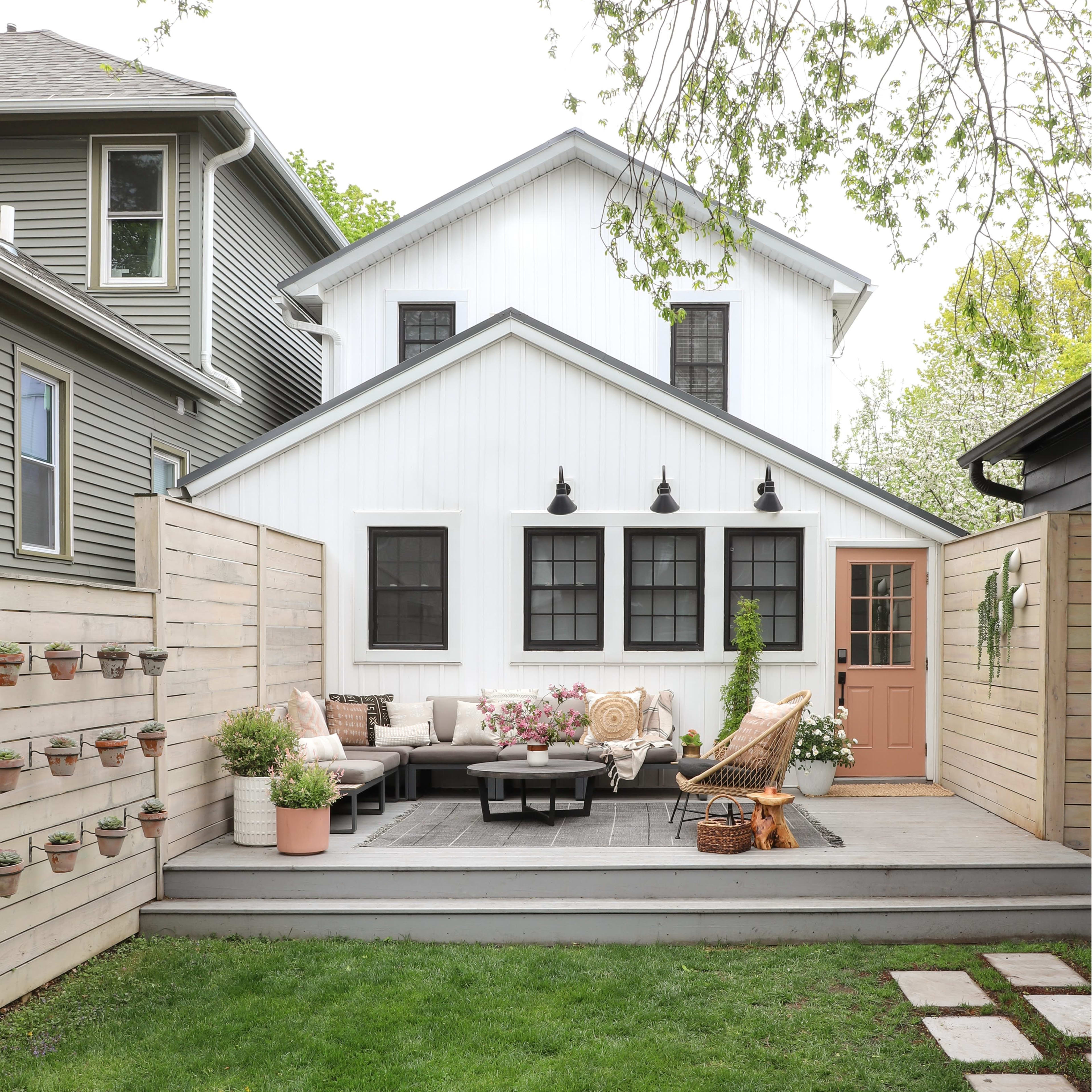 A backyard with a wooden deck featuring seating, decorative plants, and a house with a white exterior and dark windows in the background.