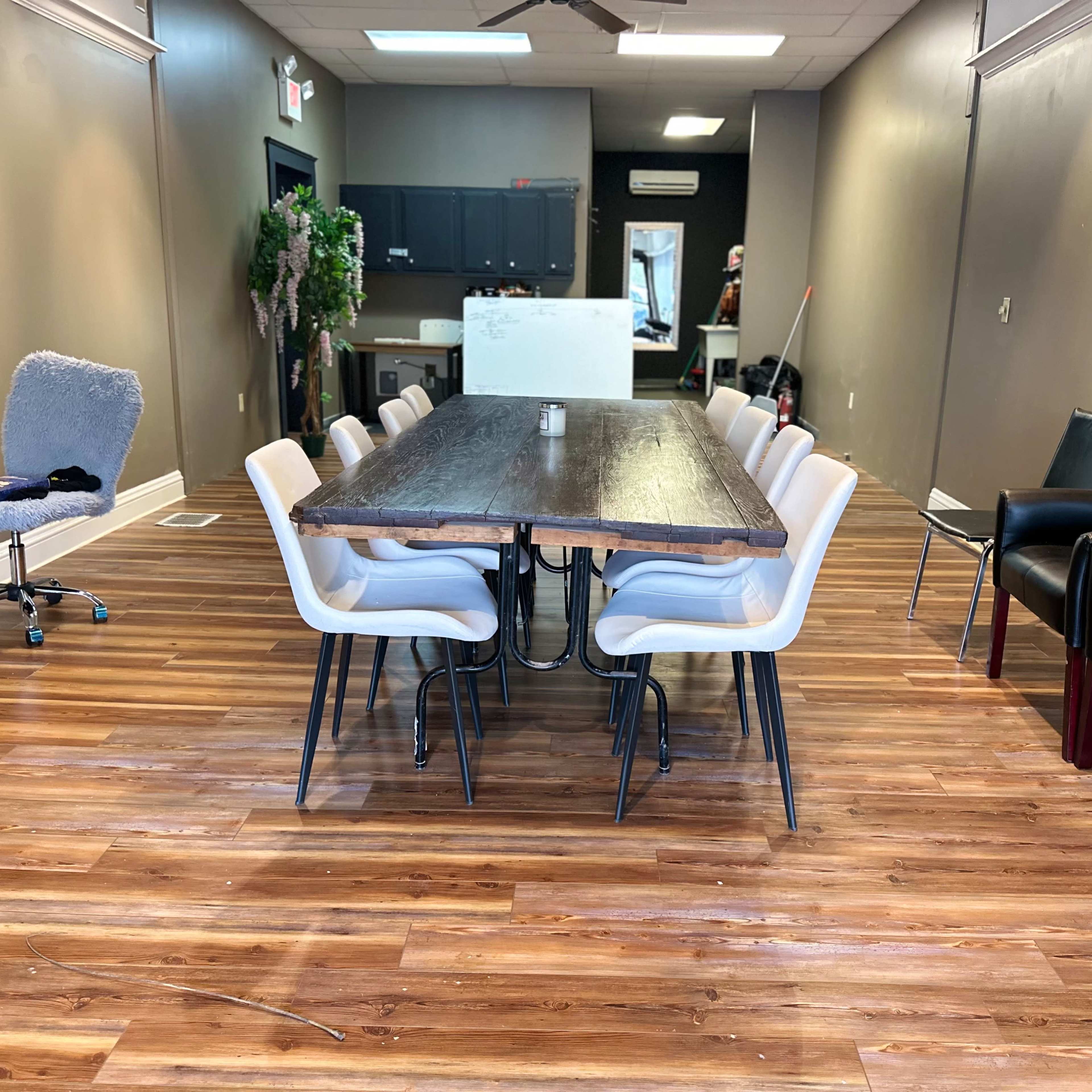 A long wooden table with white chairs is positioned in a spacious, well-lit room featuring wooden flooring and gray walls.