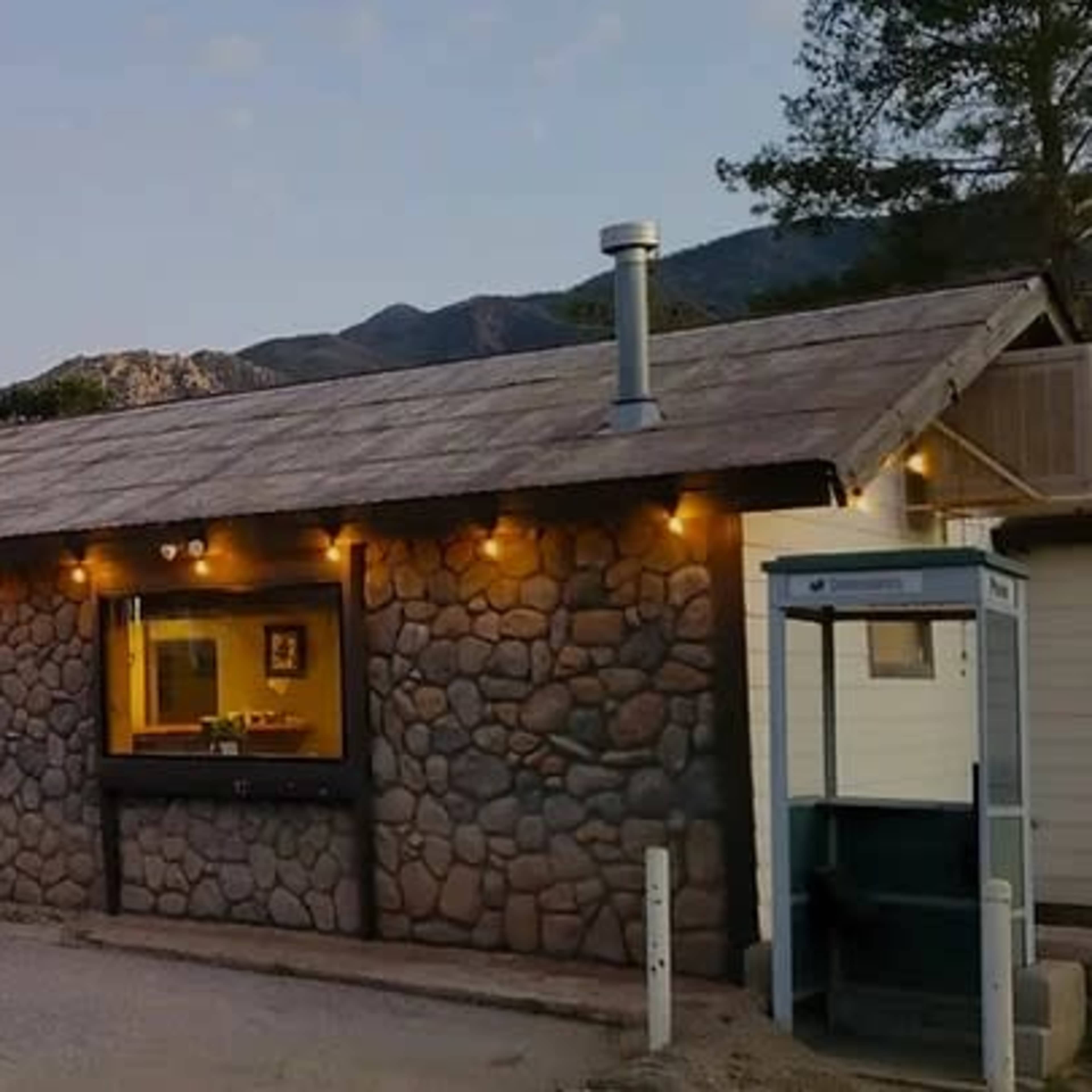 A stone building with a metal roof and string lights is shown alongside a parked black convertible car and an American flag.
