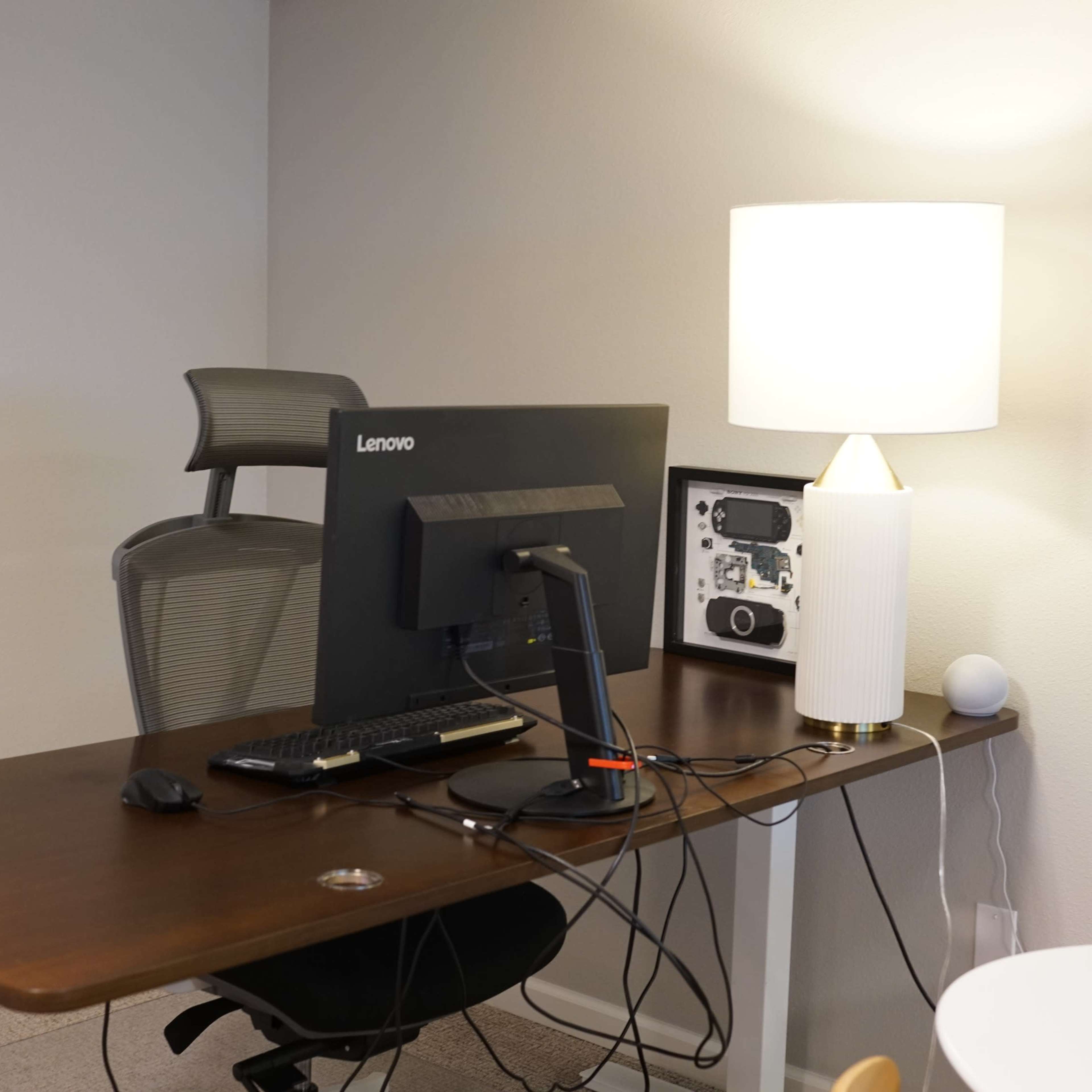 An office space features a wooden desk with a Lenovo computer, a lamp, and a chair, situated against a neutral wall.