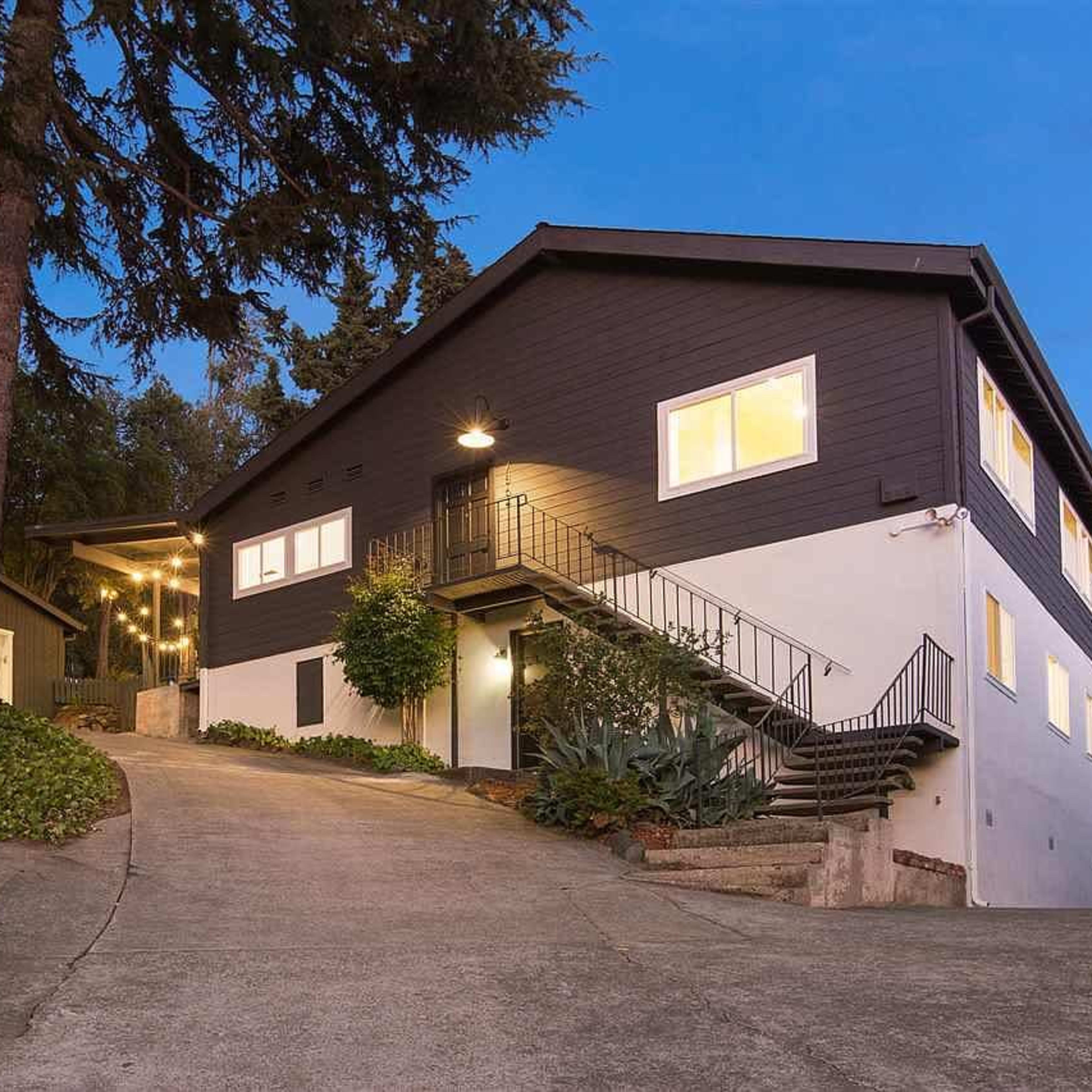 A two-story house situated on a sloped driveway, with a garage on the left and multiple windows illuminated in the evening light.