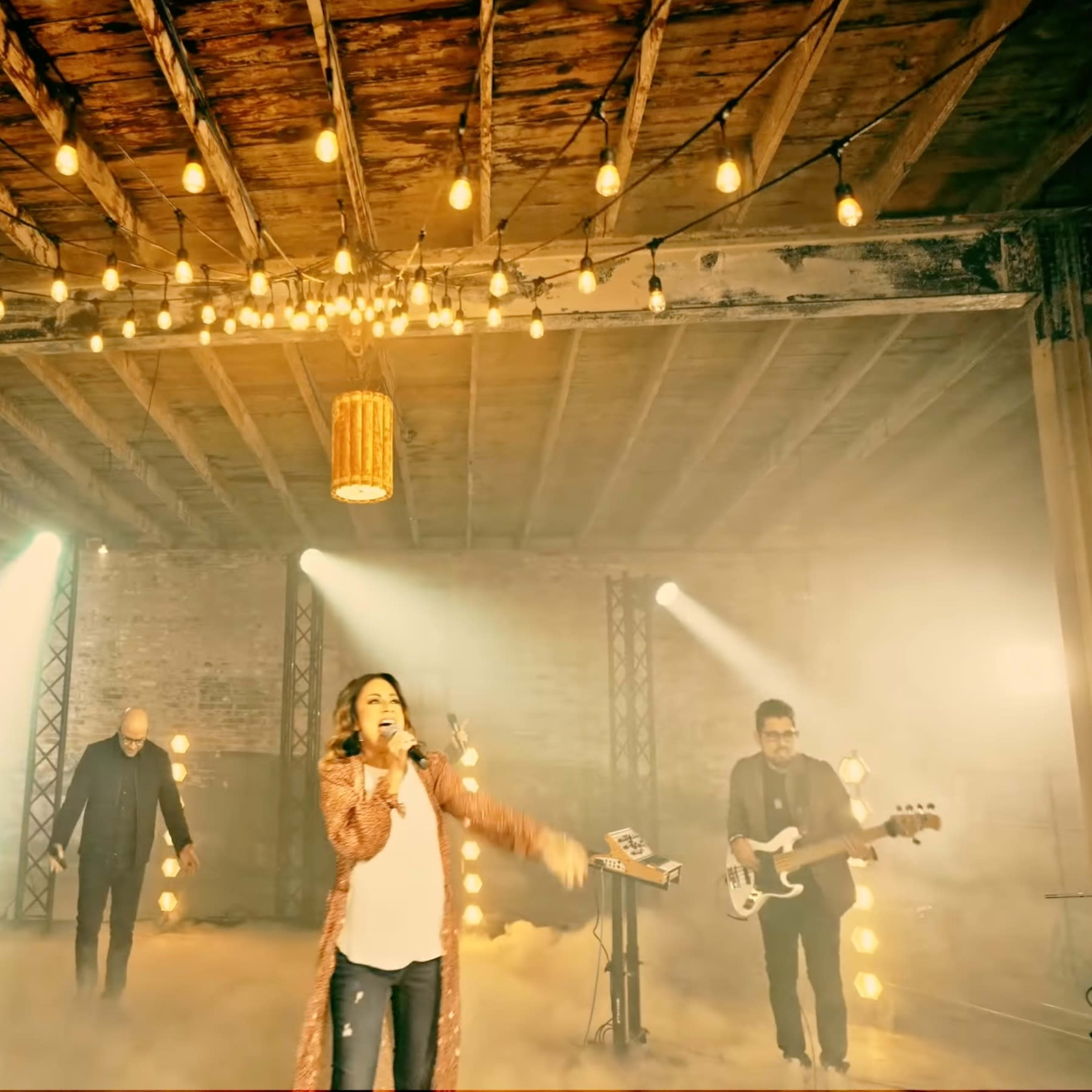 A band performs on a stage with string lights and a smoky atmosphere in a rustic setting.