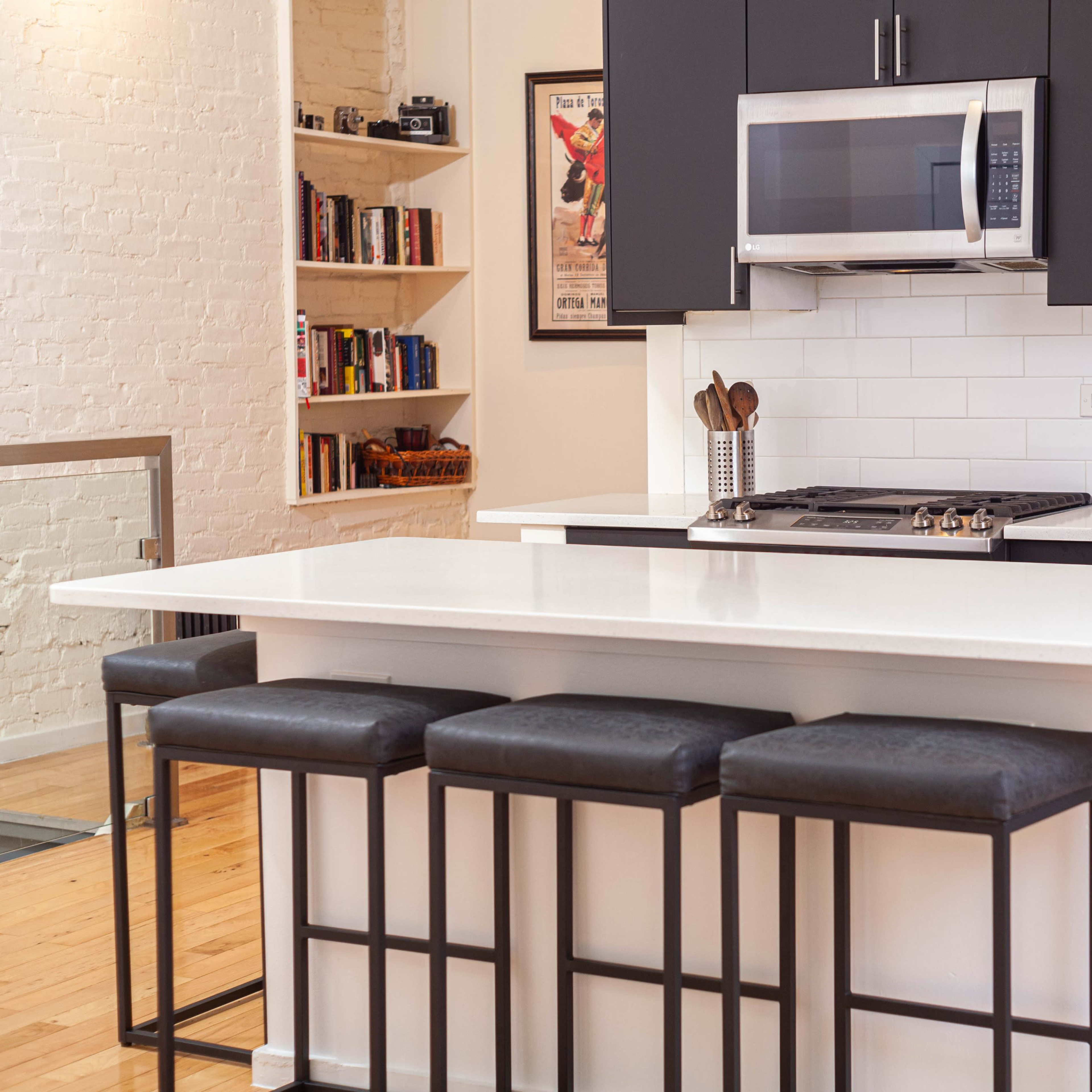 A modern kitchen featuring a white island with three black stools, stainless steel appliances, and dark cabinetry.