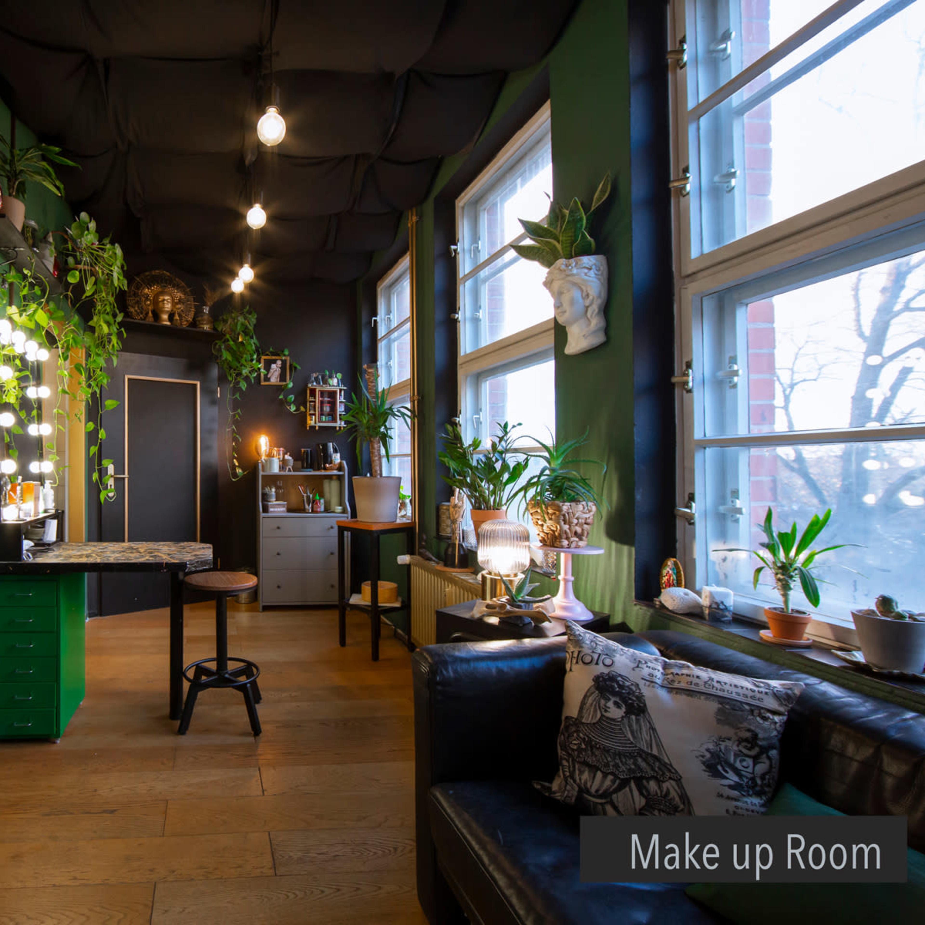A makeup room featuring a dark green color scheme, a wooden floor, and multiple plants, with a large mirror framed by lights on one wall.
