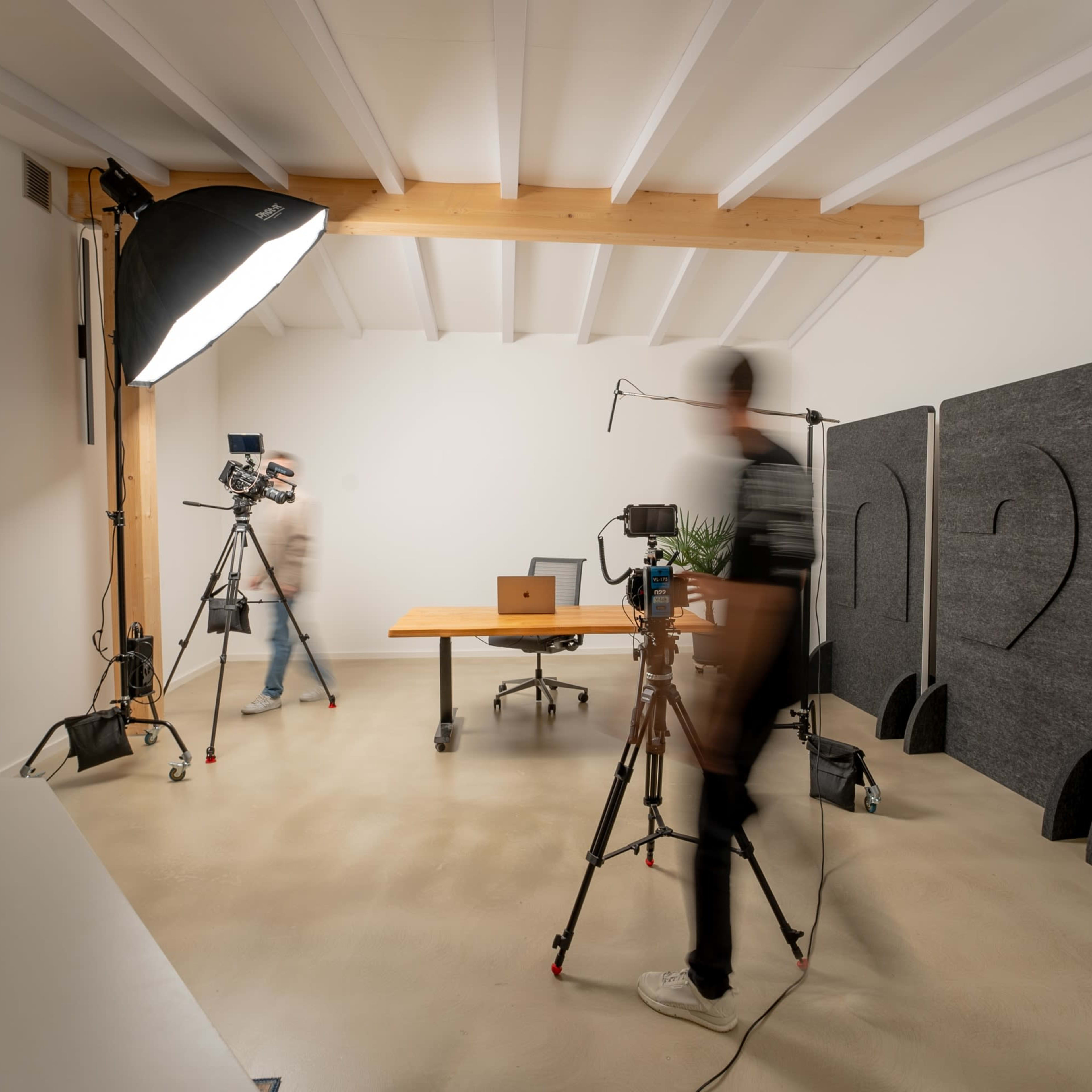 A minimalistic studio setup features a table with a laptop, two cameras on tripods, and softbox lighting in a room with wooden beams and padded soundproofing on the walls.