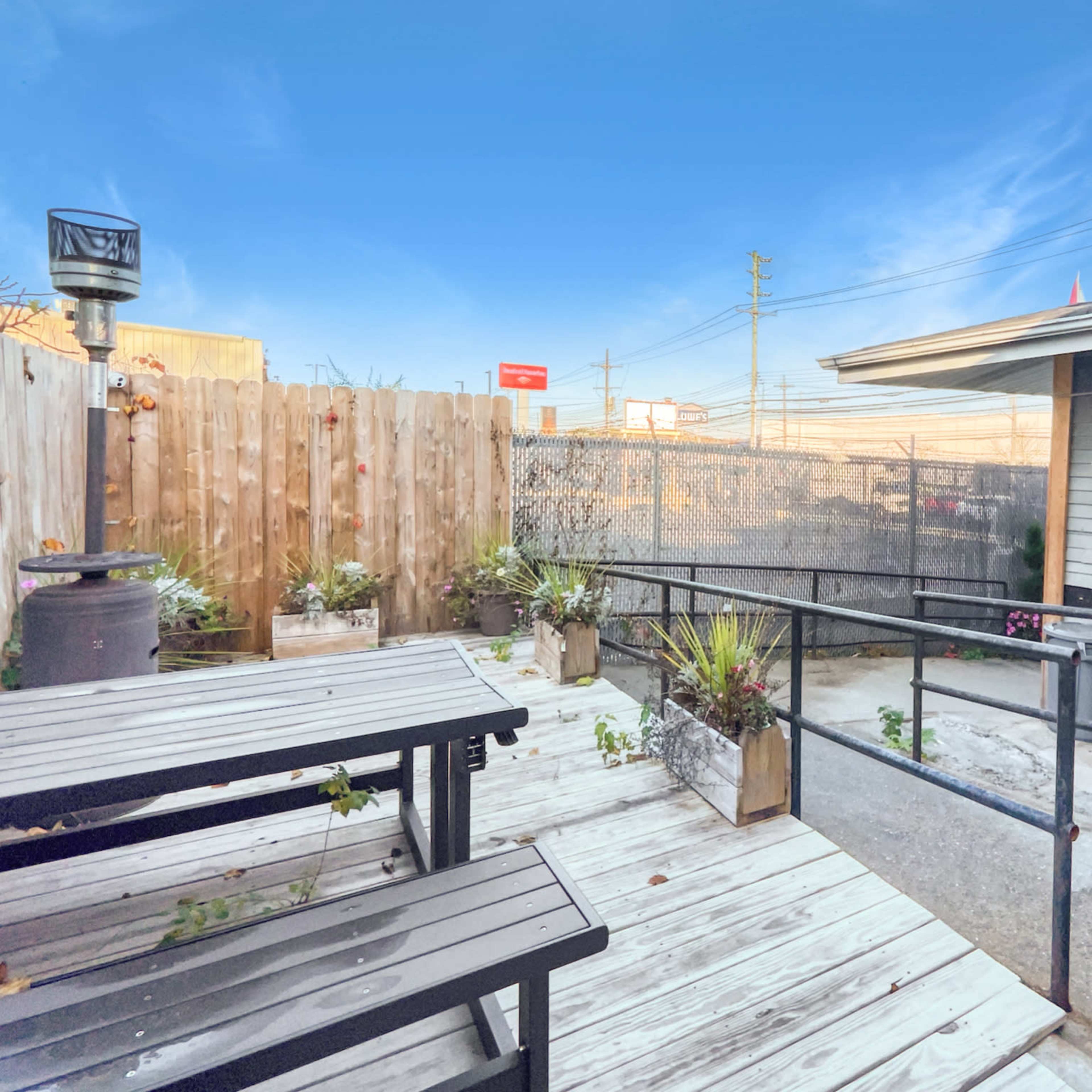 A wooden deck area with two benches, potted plants, and a fenced enclosure, next to a building under a clear blue sky.