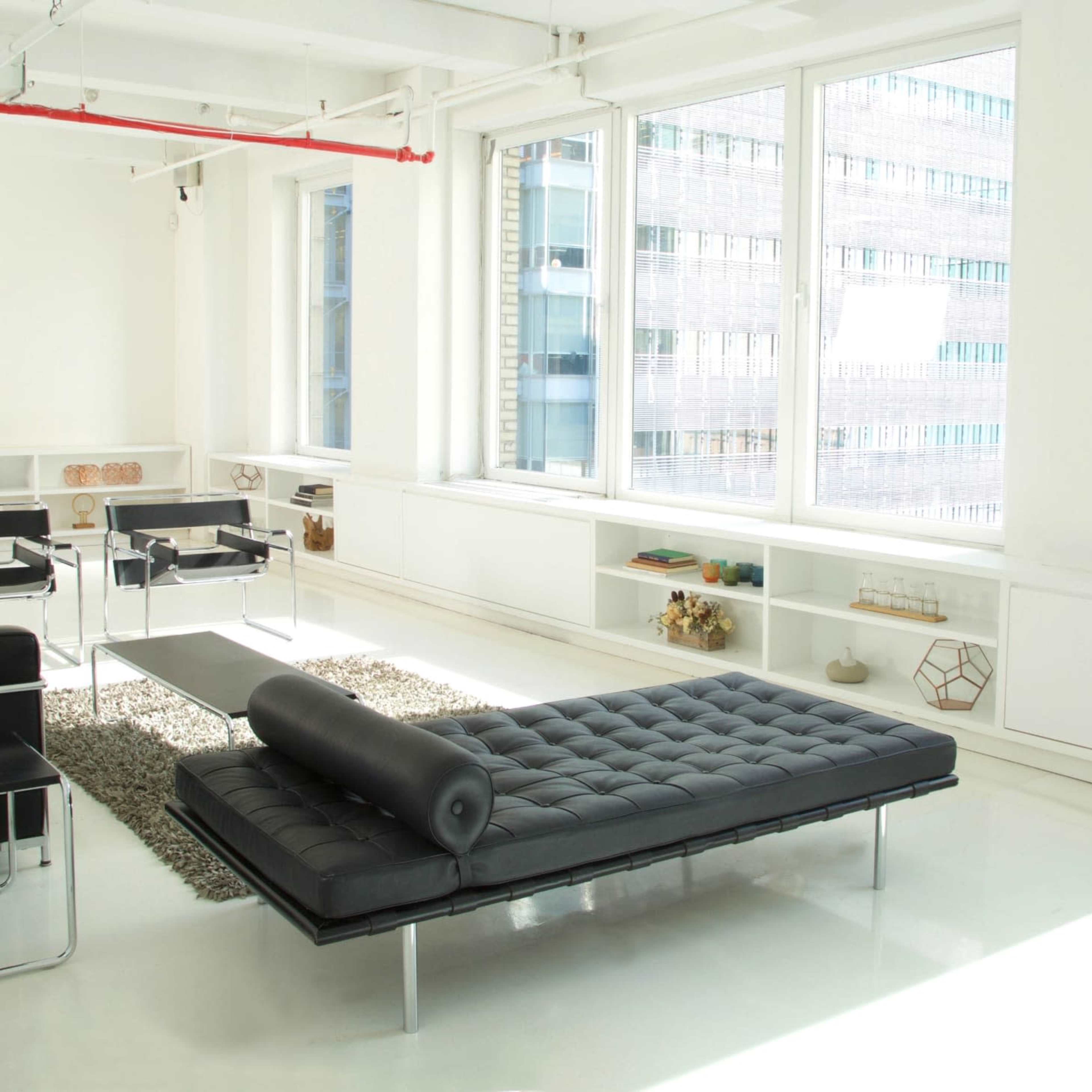 A modern, minimalist office space with large windows, a black lounge chair, and a tufted daybed positioned on a rug.