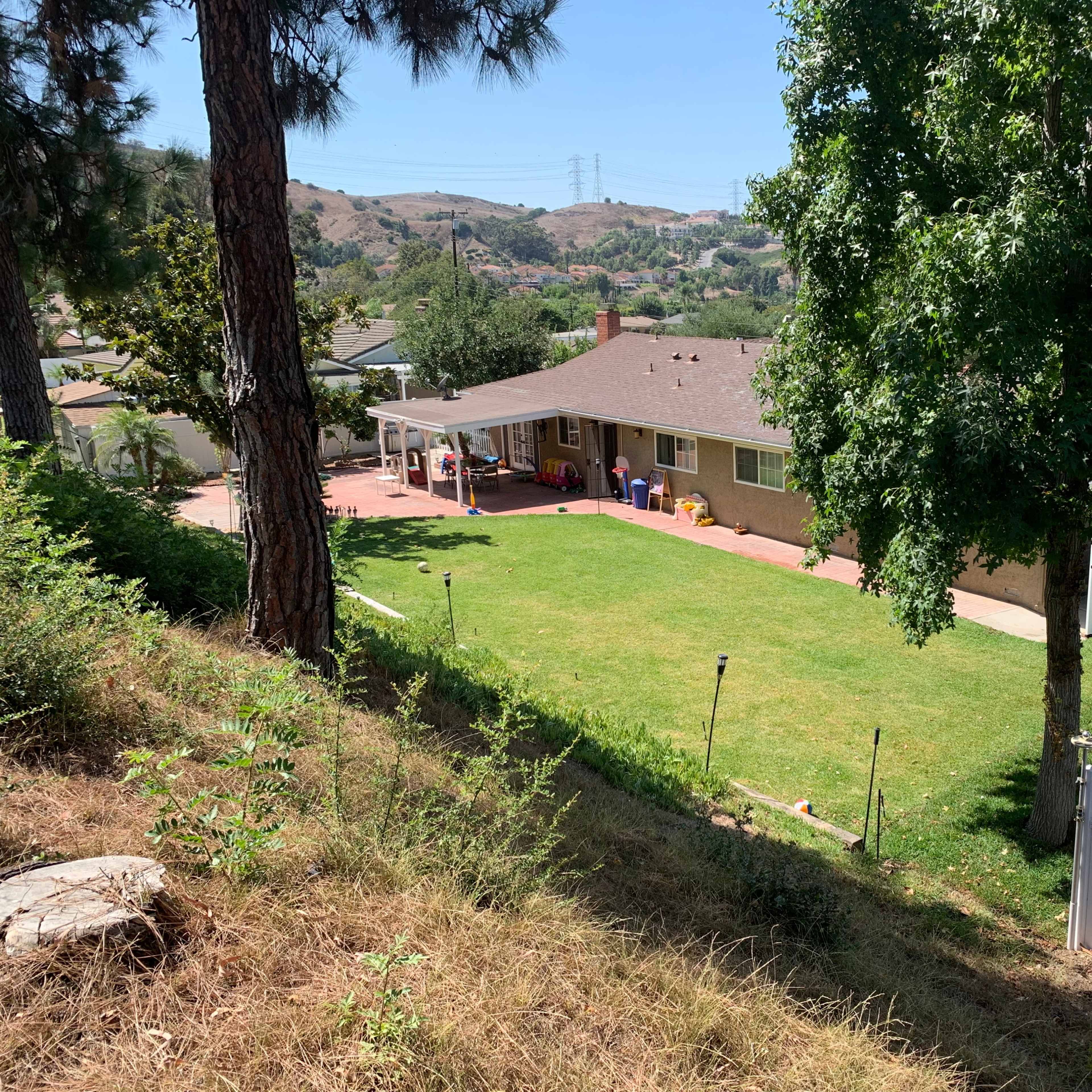 A single-story house with a shaded backyard and grassy area is surrounded by trees and hills.