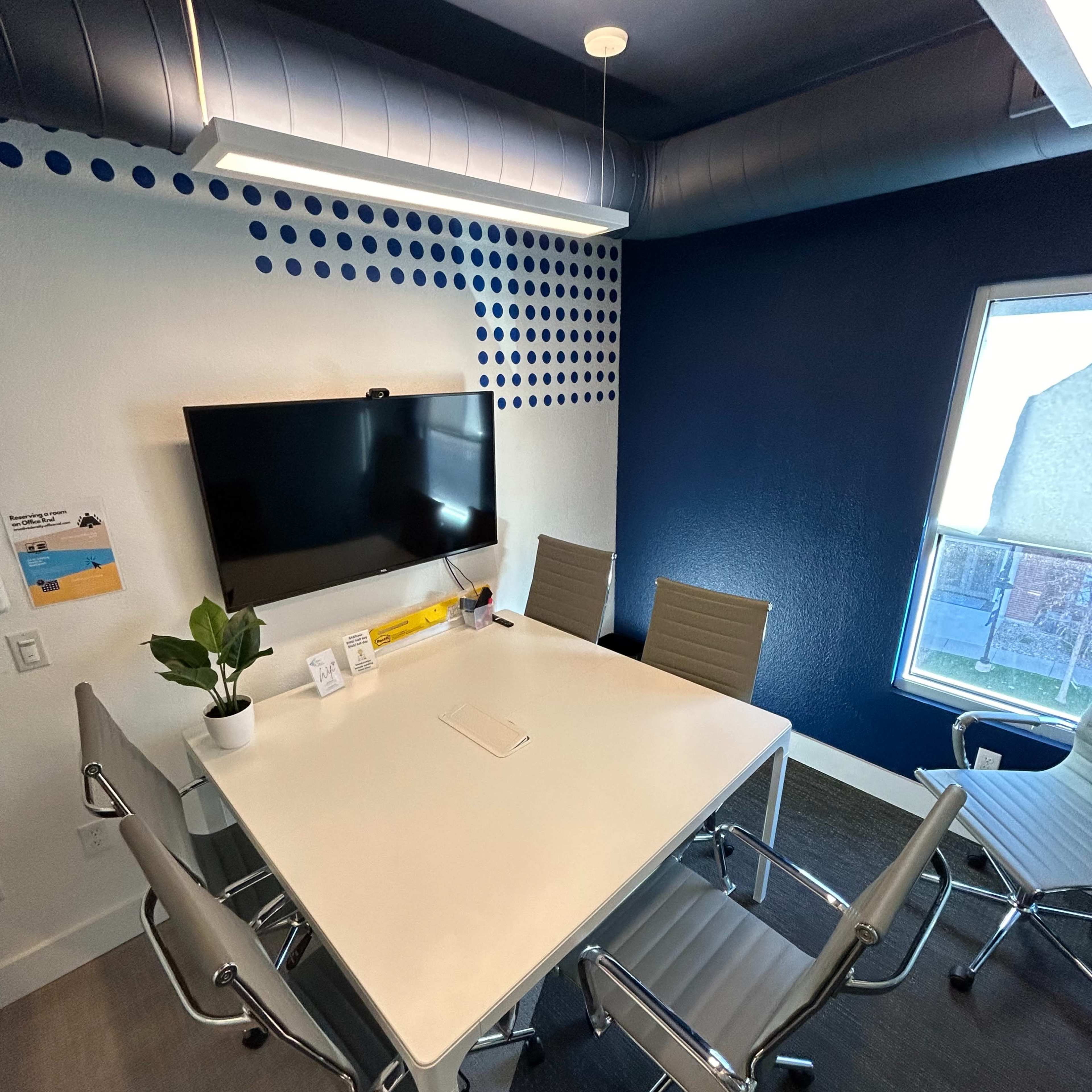A small conference room with a white table, four metal chairs, a wall-mounted television, and a window.