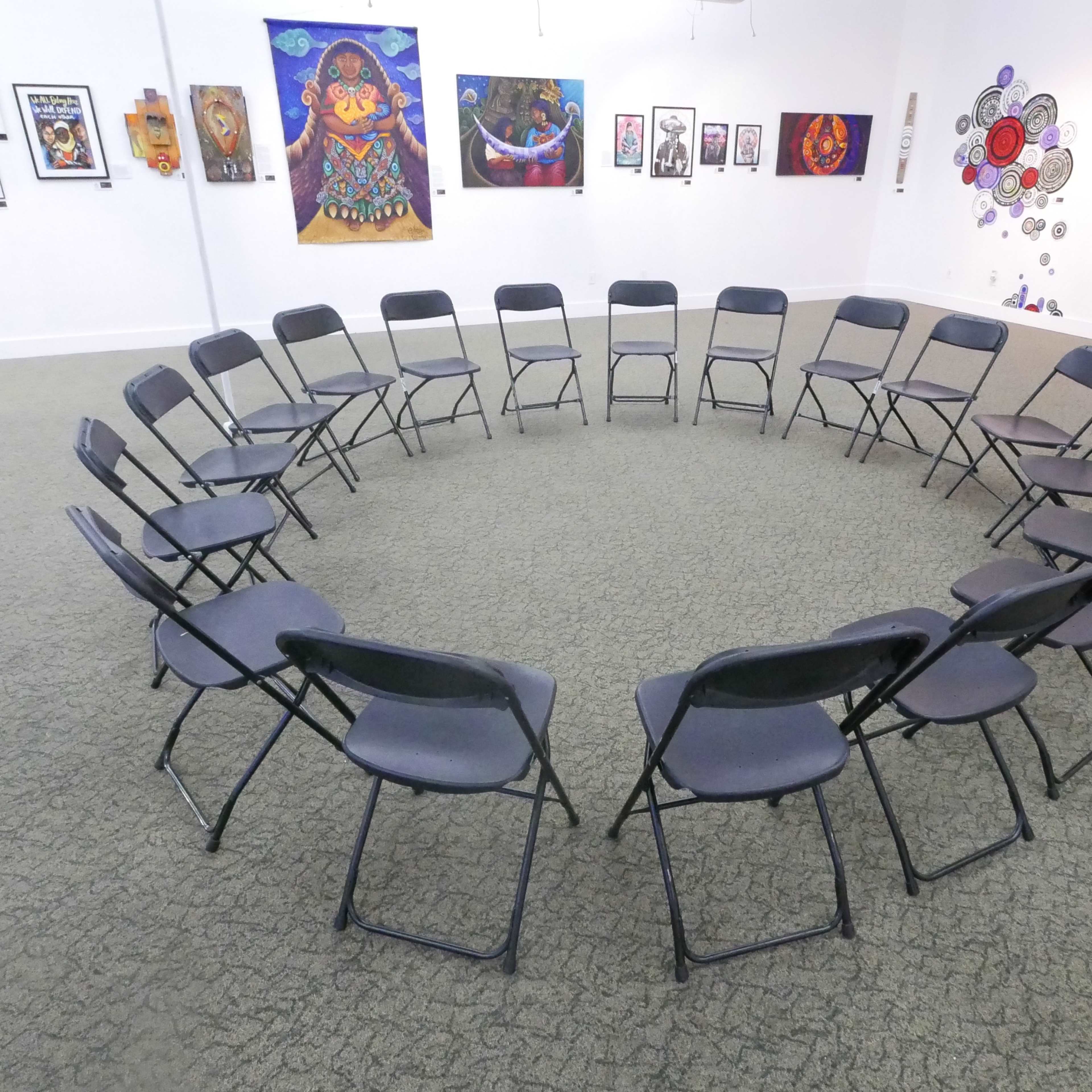 A circular arrangement of black folding chairs is set up in an art gallery adorned with various colorful artworks on the walls.