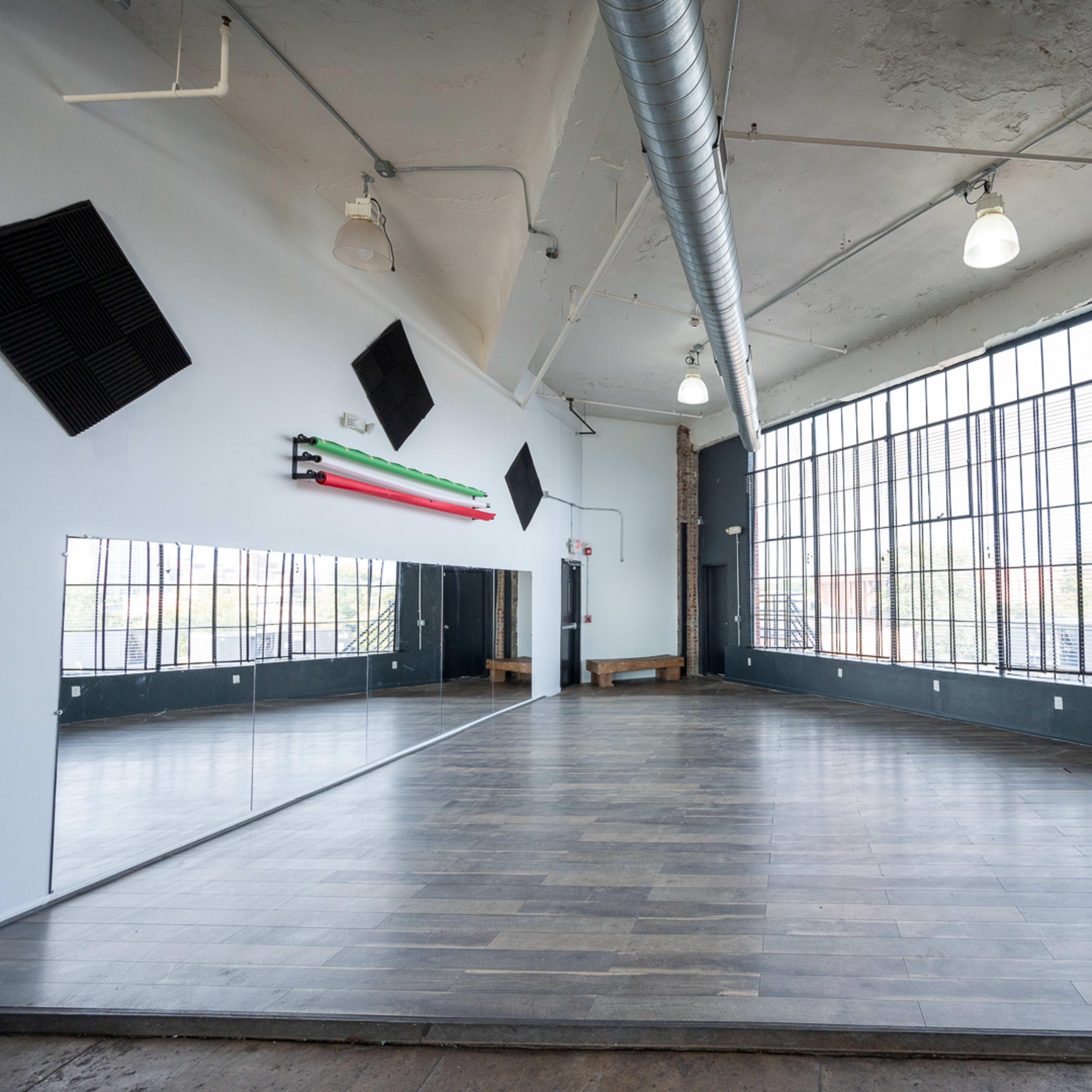 A spacious, well-lit room features large windows, wooden flooring, and a mirror along one wall.