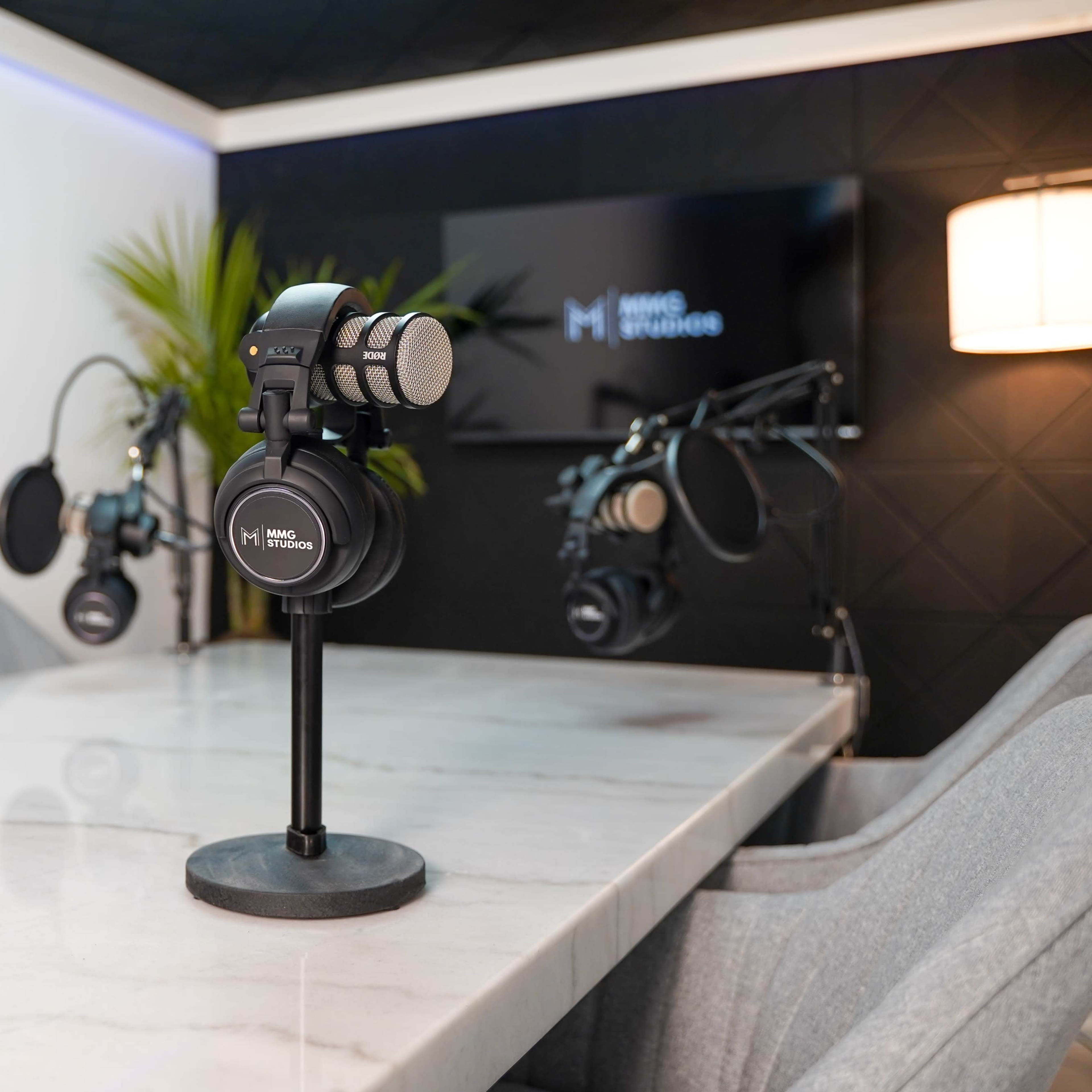 A modern podcast studio features multiple microphones on stands, headphones, and a sleek marble table against a dark wall with a television.