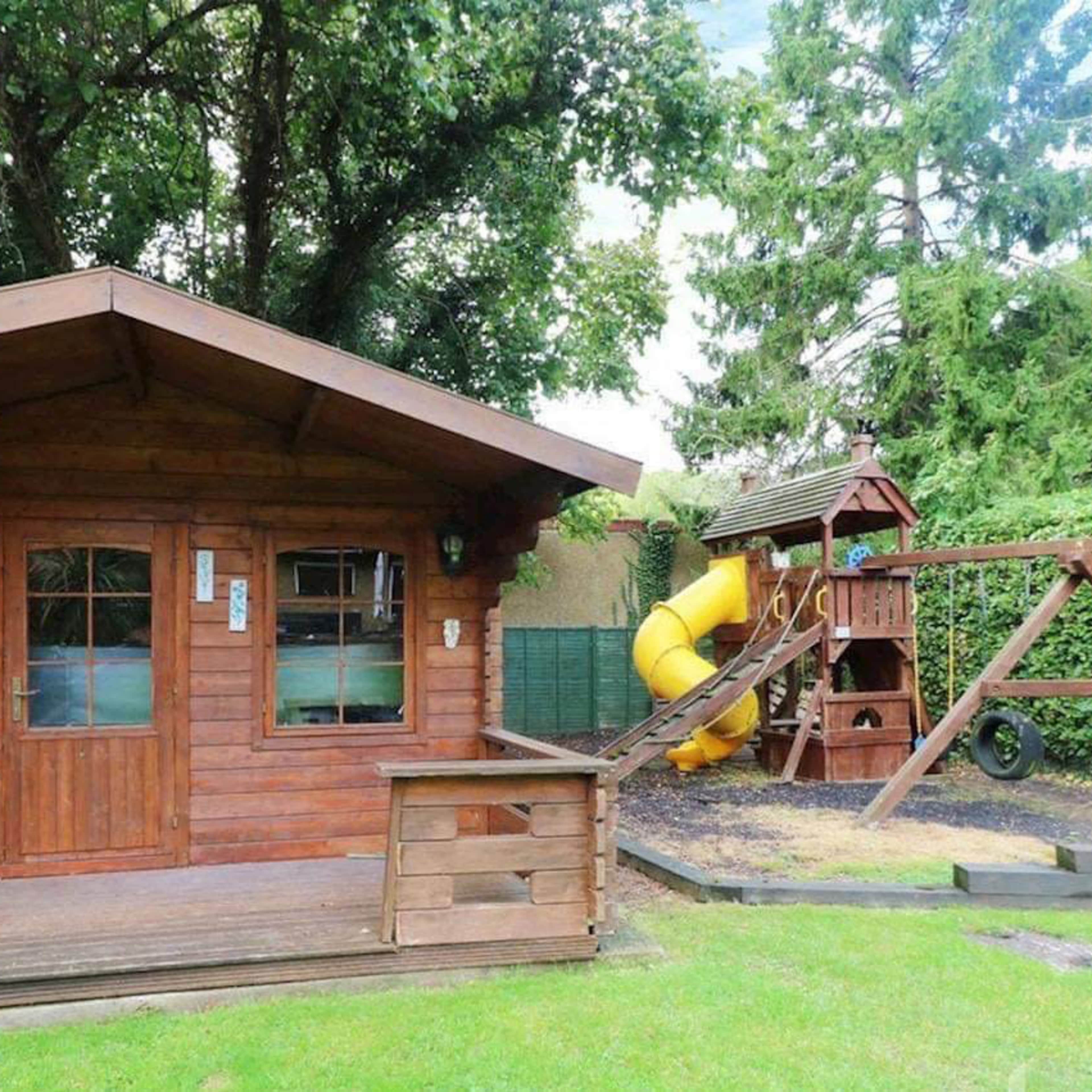 A wooden garden cabin with a porch stands next to a playset featuring a slide and swing in a grassy yard.