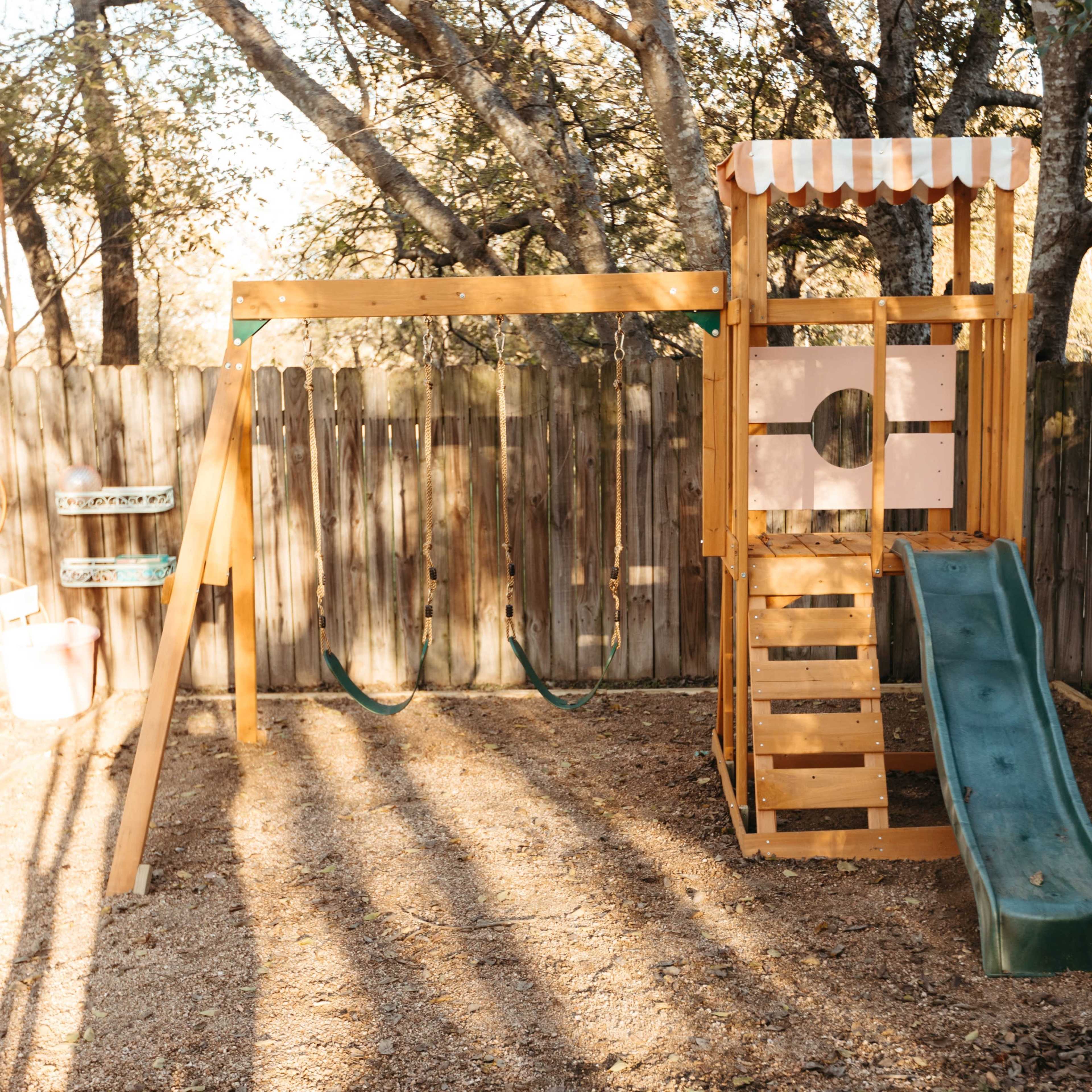 A wooden playset with a slide and swings is located in a yard surrounded by a wooden fence and trees.