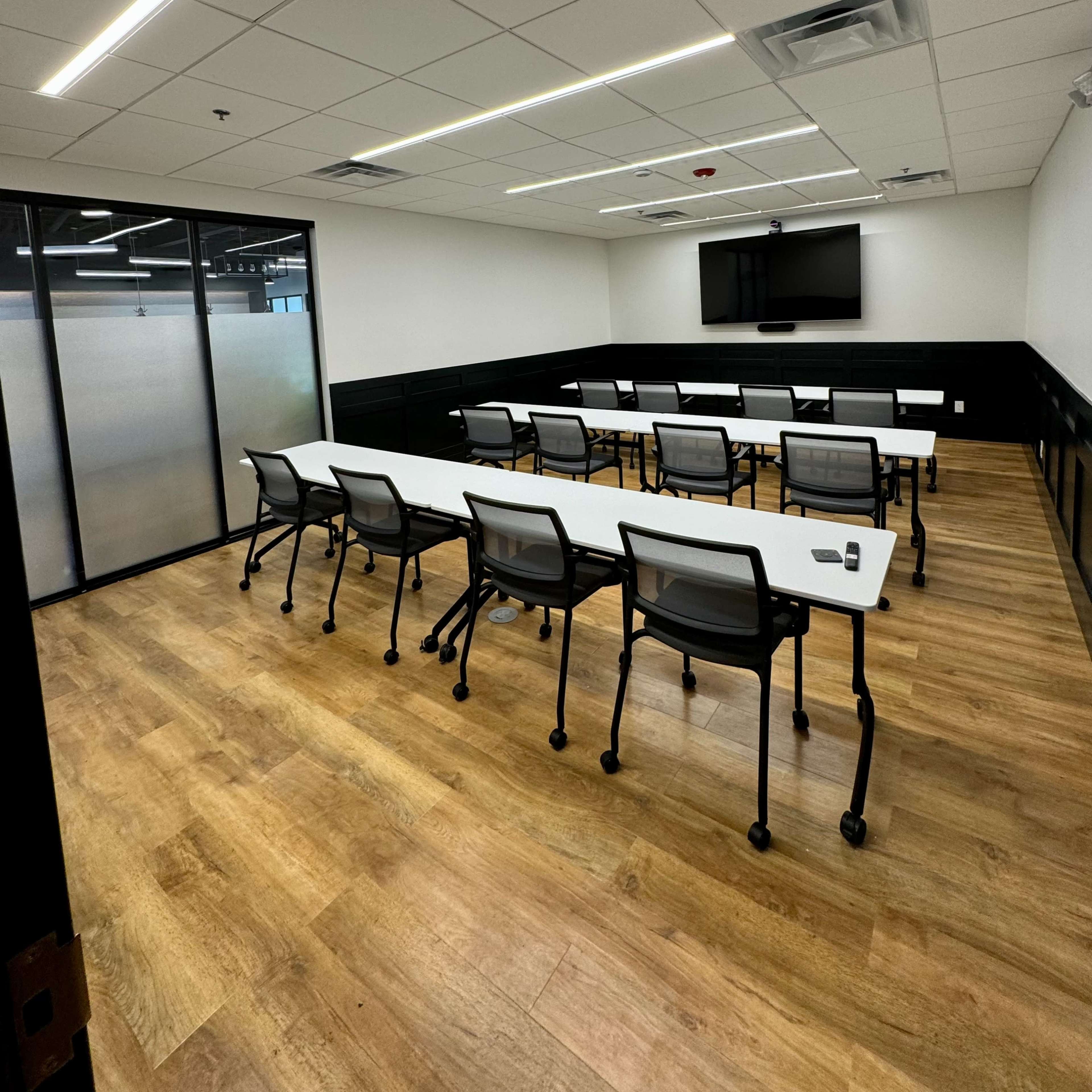 A modern conference room with rows of tables and chairs facing a flat-screen TV on the wall.