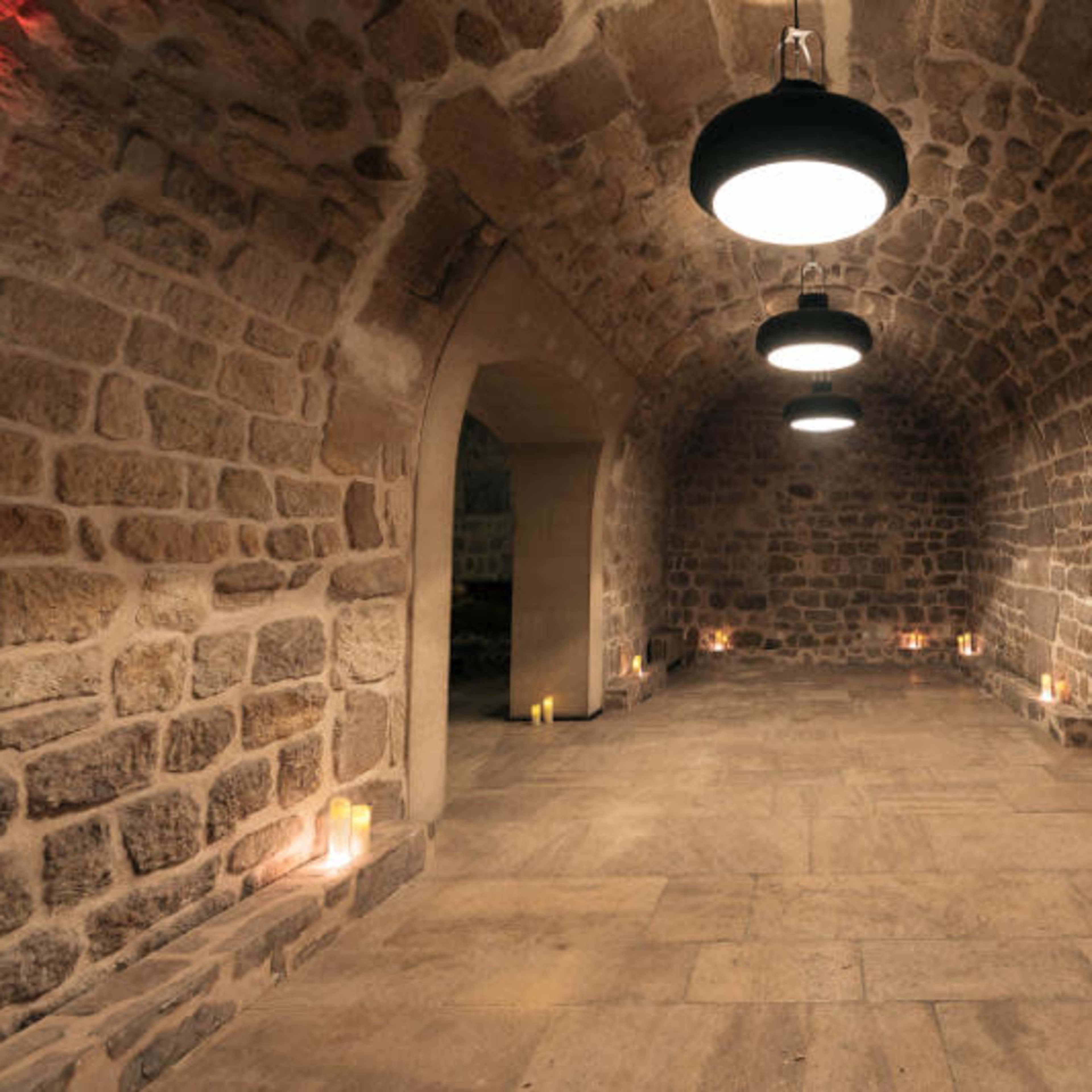 A stone-walled room with three hanging lights and candles placed along the perimeter of the floor.