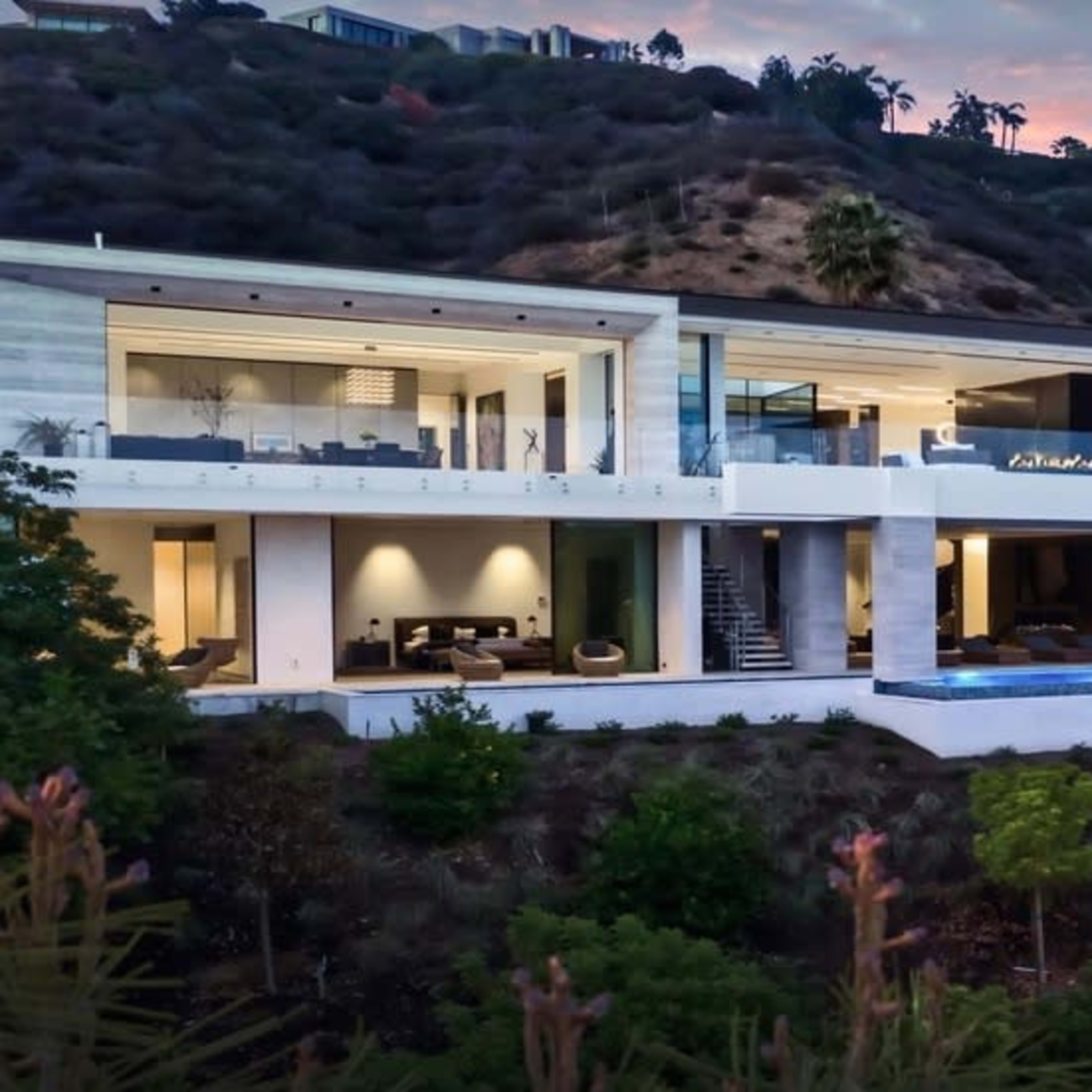A modern hillside mansion features multiple levels, large glass windows, and an outdoor pool surrounded by lush greenery.