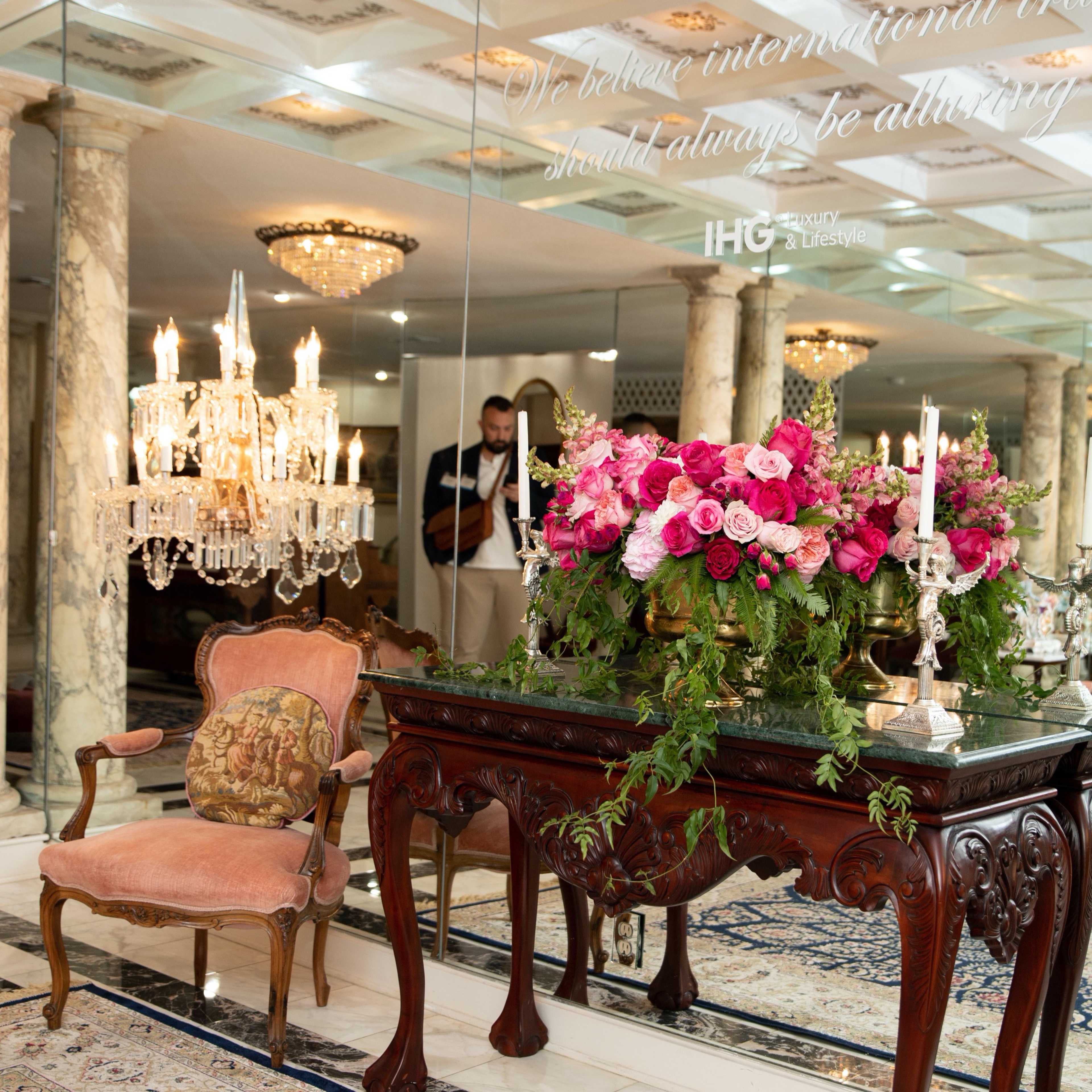 A table adorned with a vibrant floral arrangement of pink roses is positioned in front of a mirrored wall in an elegantly decorated room featuring chandeliers and ornate furnishings.