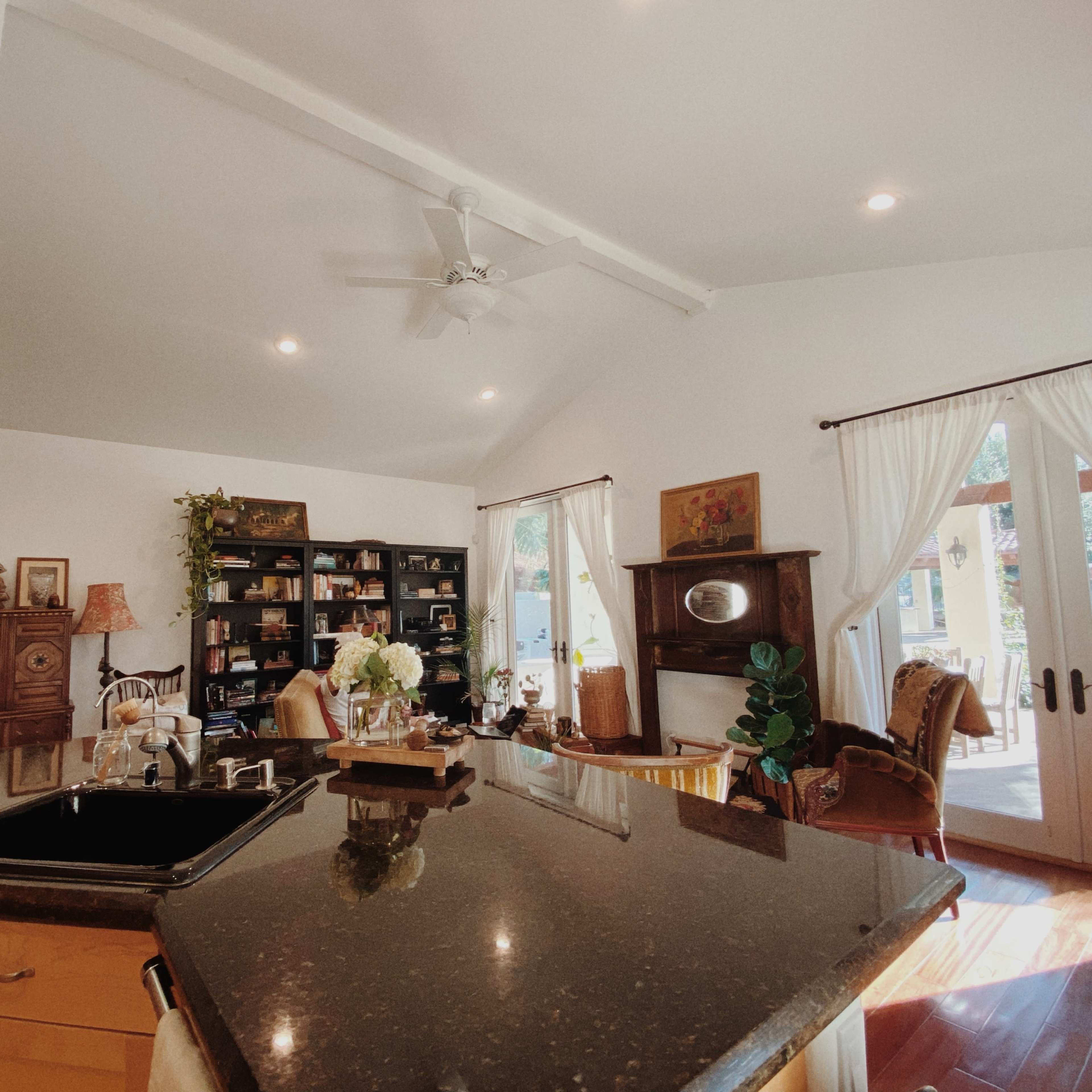 A bright kitchen with wooden cabinets, a black countertop, and an adjoining living area featuring bookshelves and large windows.