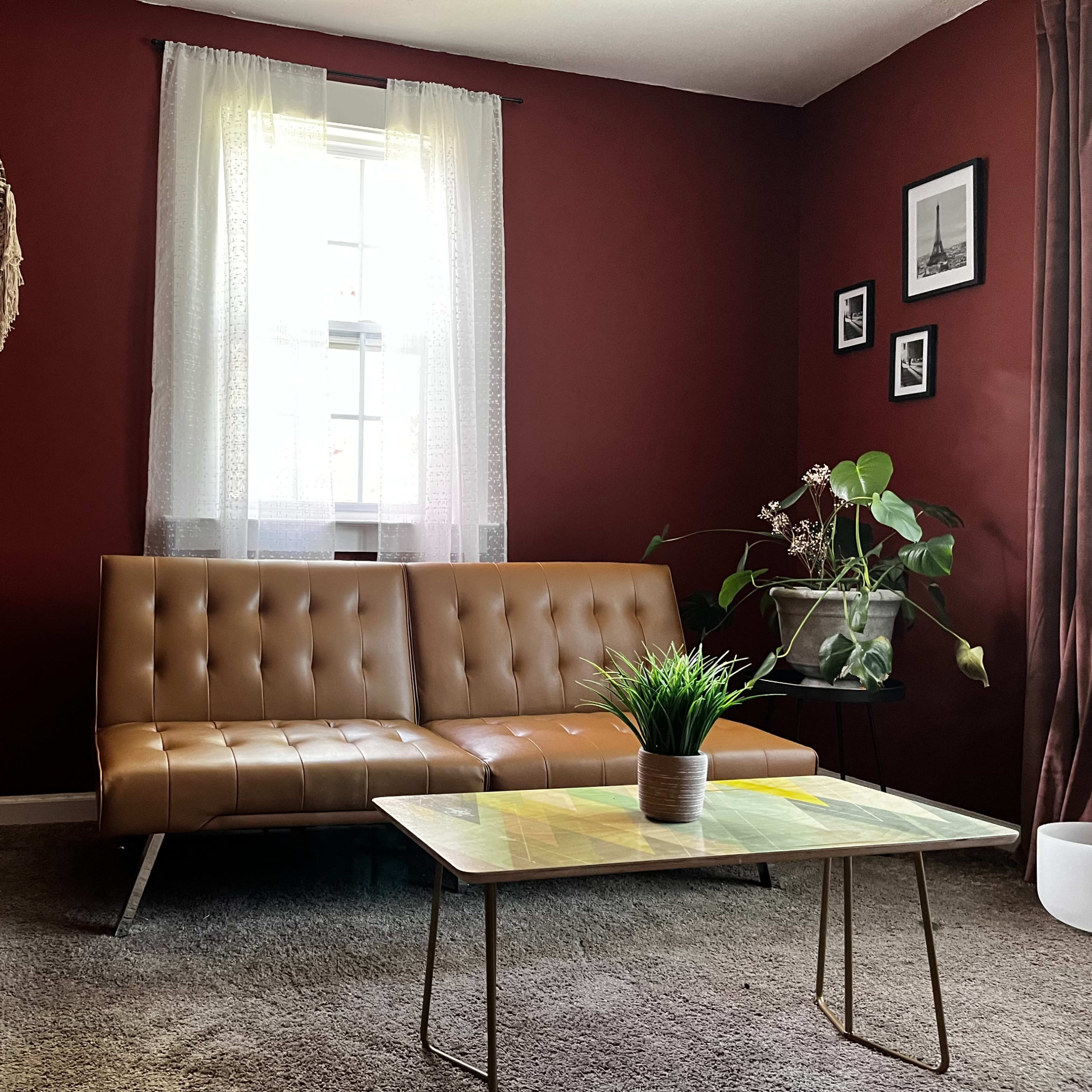 A living room with a brown tufted sofa, a glass coffee table, a potted plant, and decorative wall art against a red backdrop.