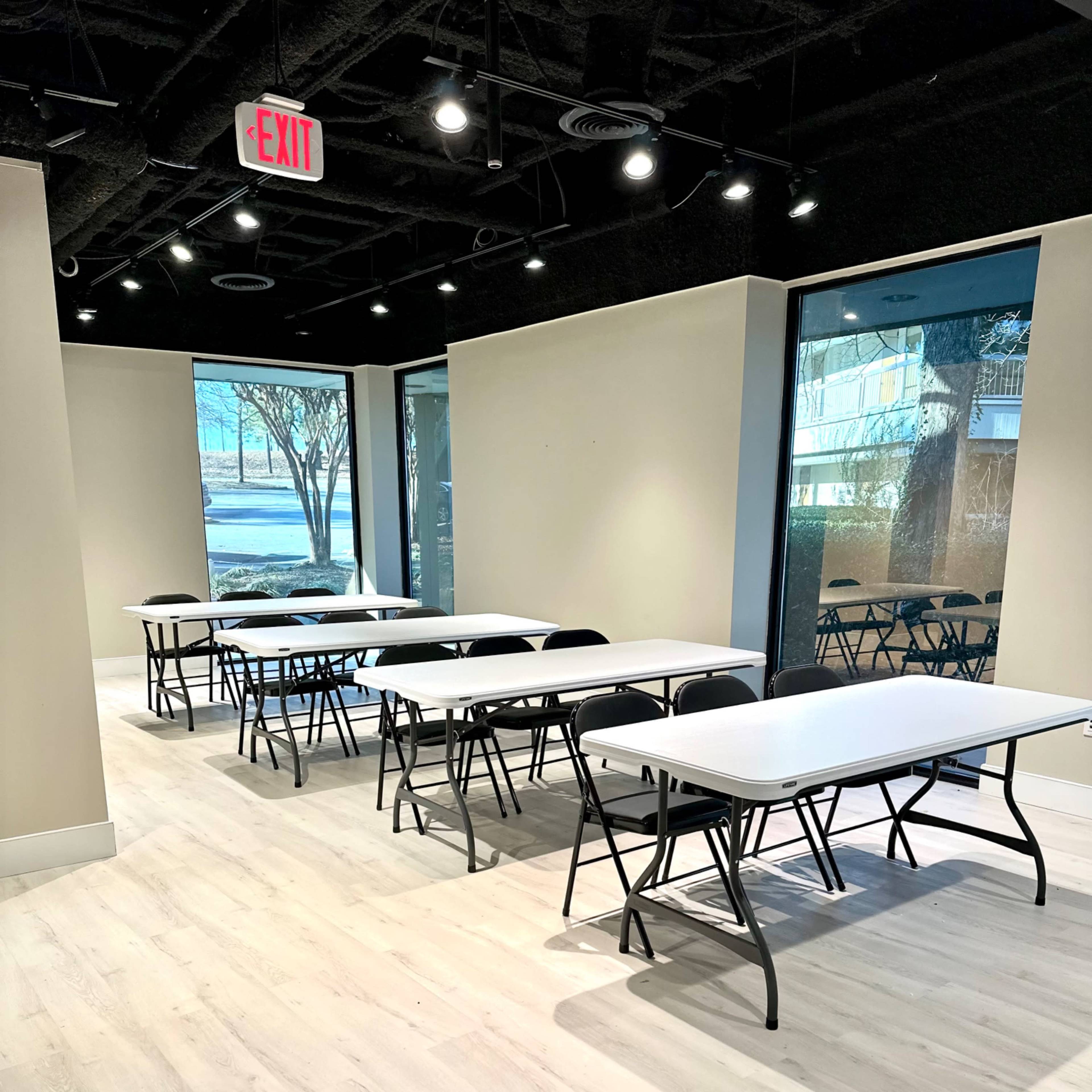 A spacious room with several rows of folding tables and chairs arranged neatly, illuminated by natural light coming through large windows.