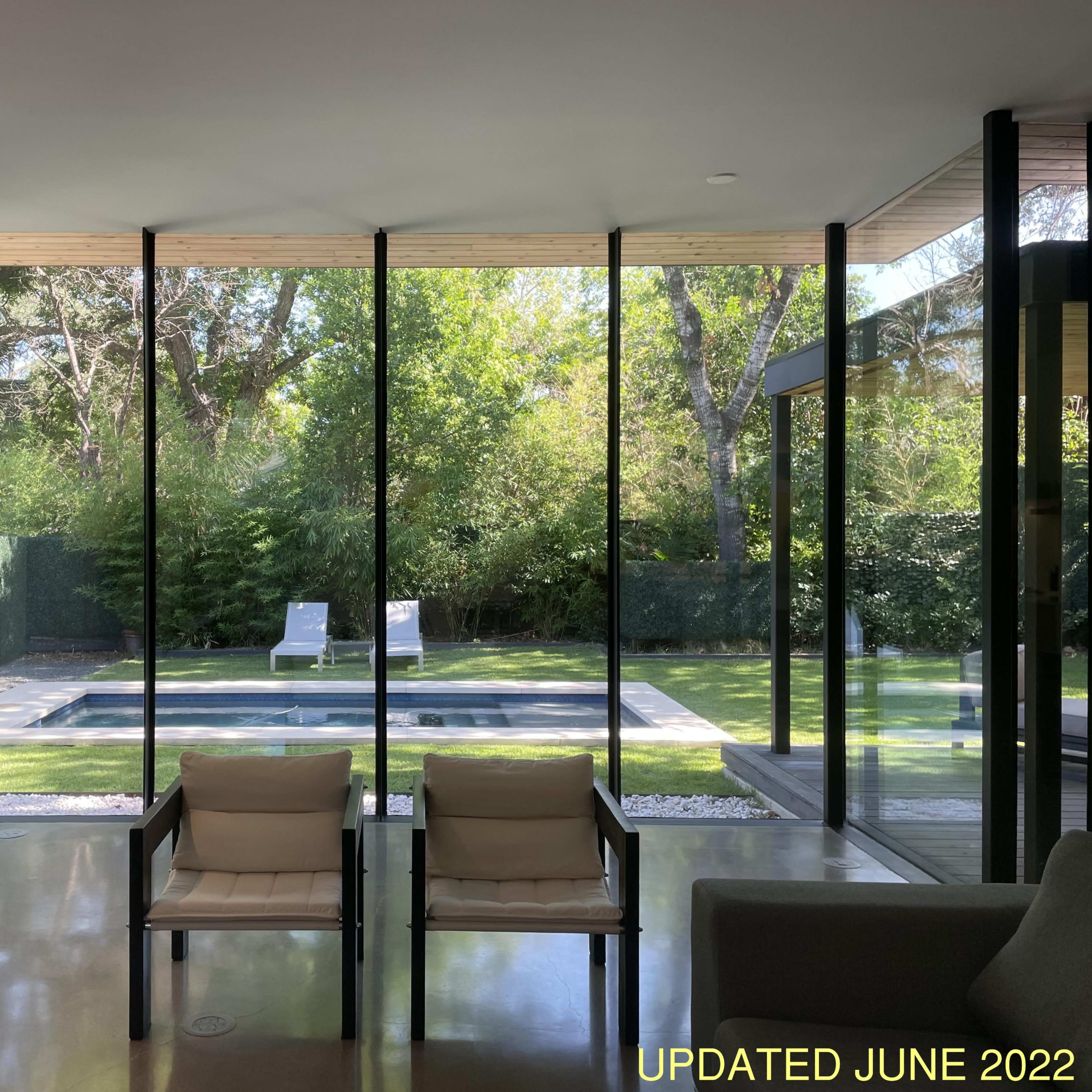 A modern interior space with two chairs facing large glass windows that overlook a garden with a pool and two lounge chairs.