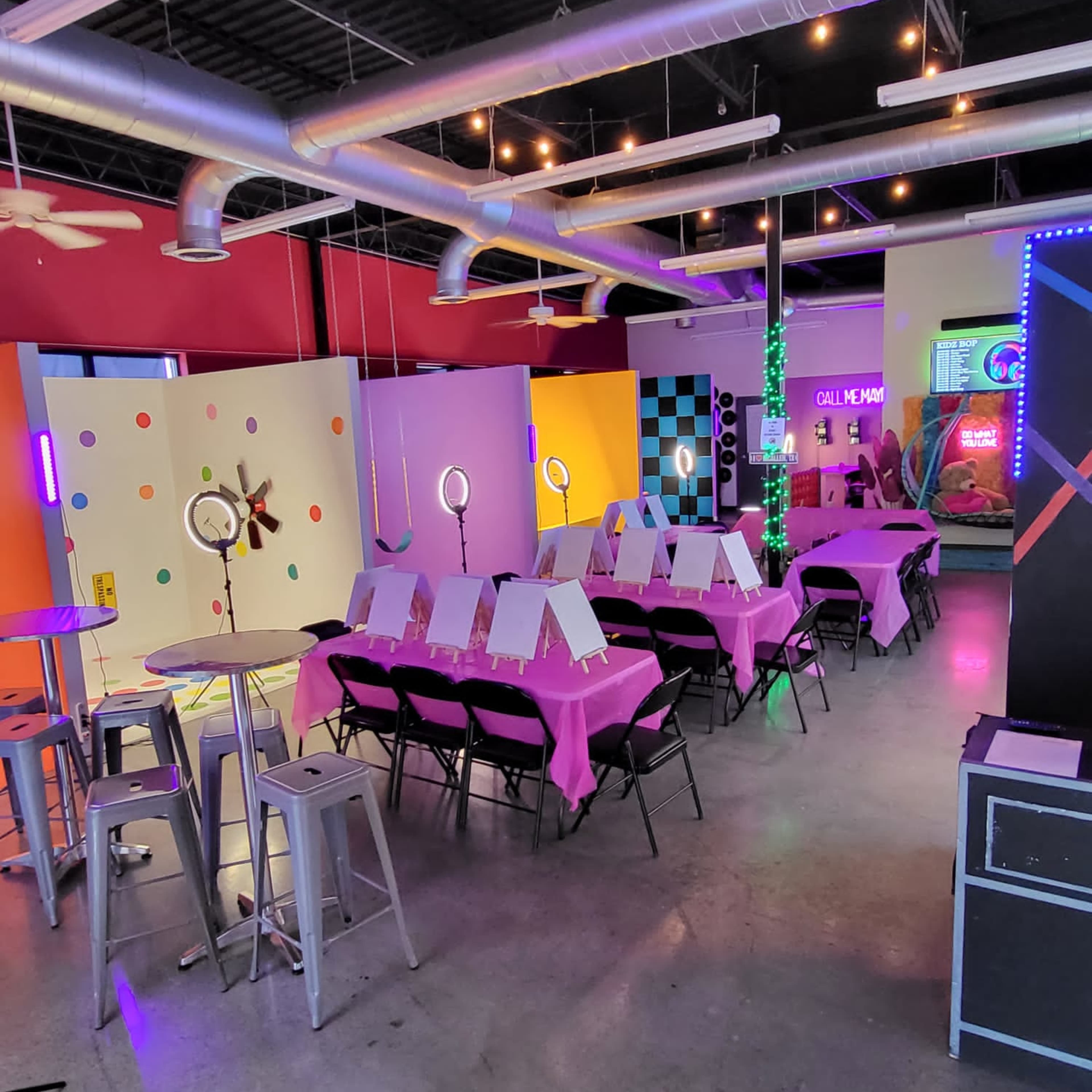 A brightly colored event space set up with tables covered in pink cloth, each featuring an easel, surrounded by colorful decorations and neon lights.