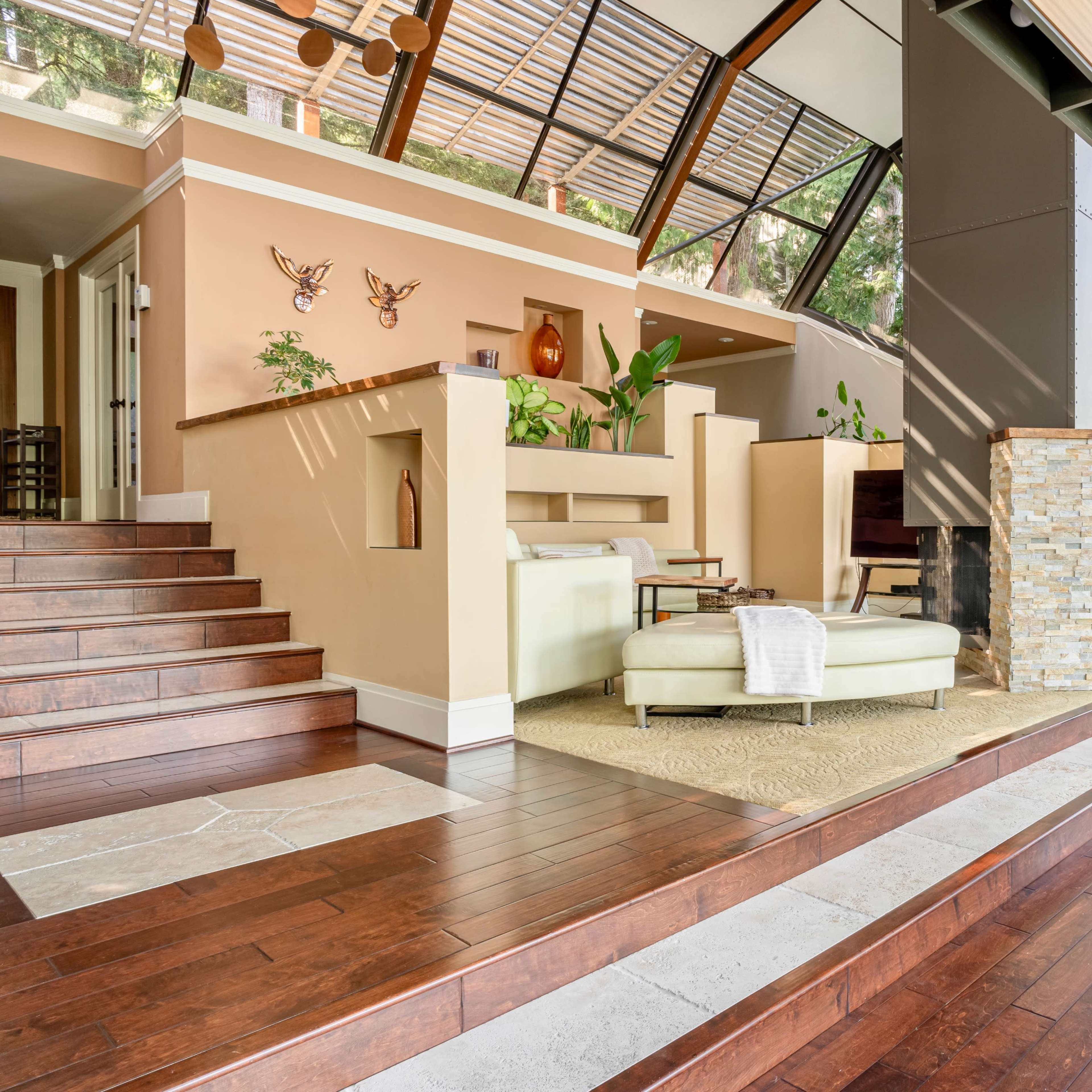 A modern living space with large glass windows, wooden flooring, and a minimalist design featuring a sectional sofa and a stone fireplace.