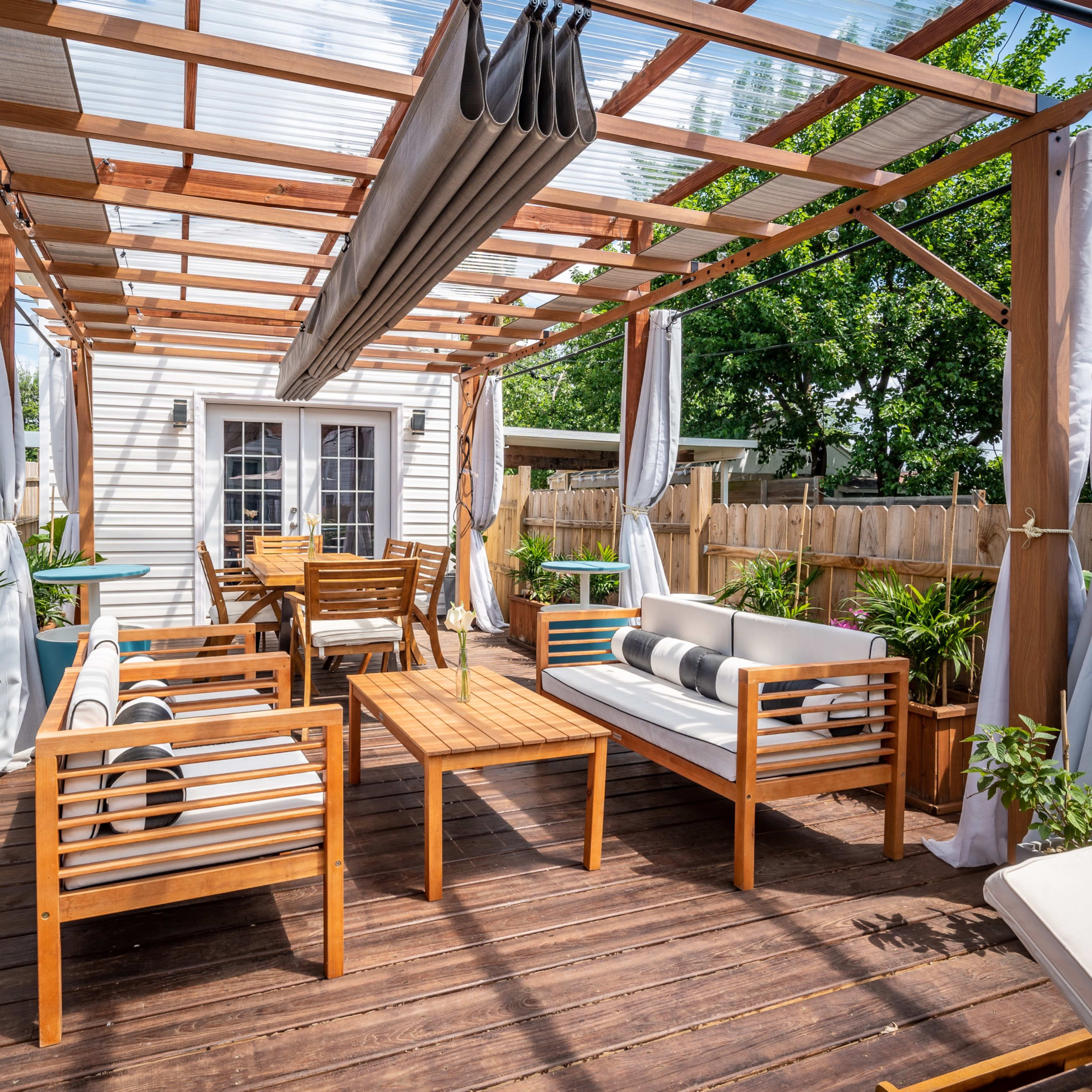 A wooden deck patio area with a pergola, featuring seating arrangements, a dining table, and potted plants.