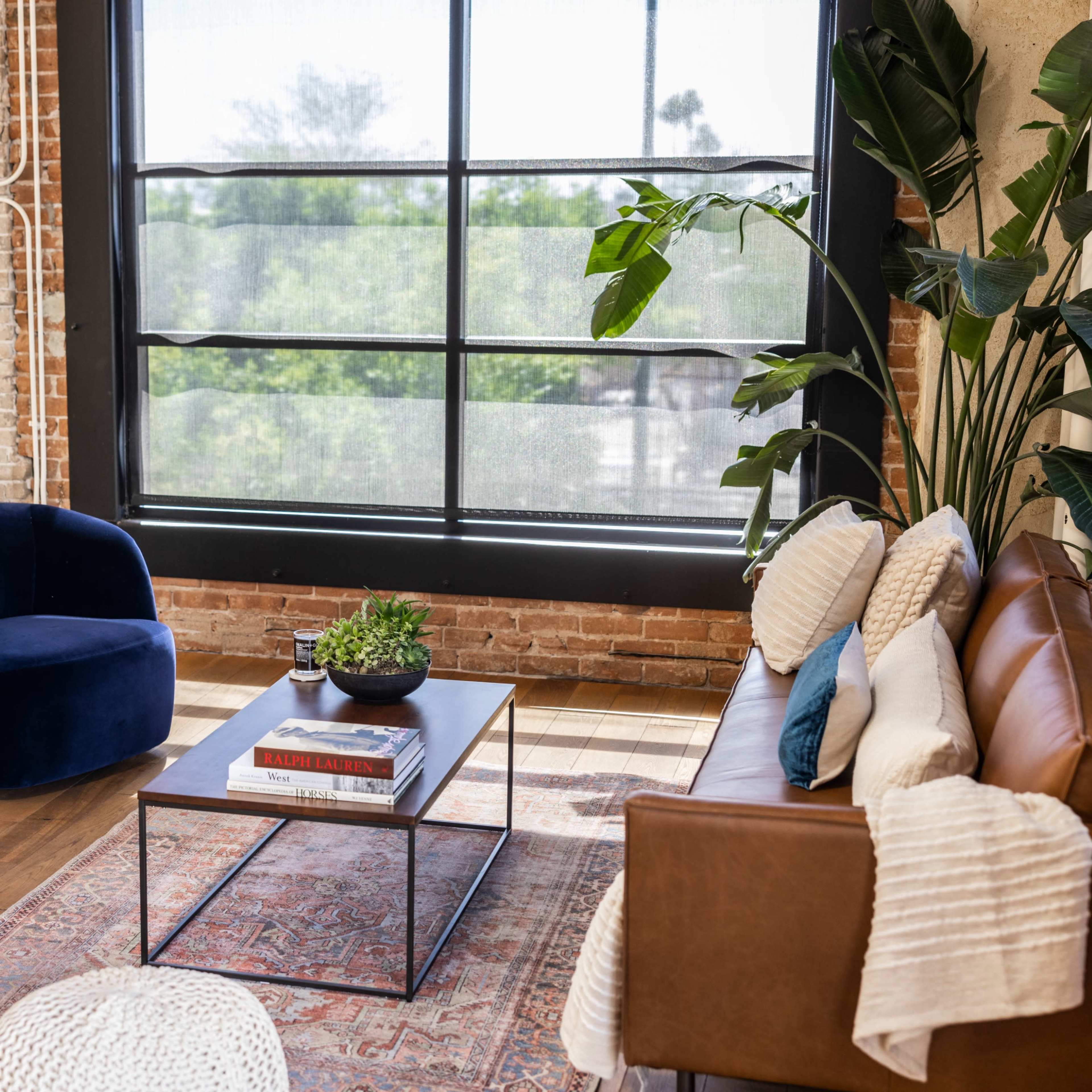 A modern living room featuring a brown leather sofa, two blue velvet chairs, a coffee table with a plant and books, and large windows allowing natural light to enter, set against a backdrop of exposed brick walls.