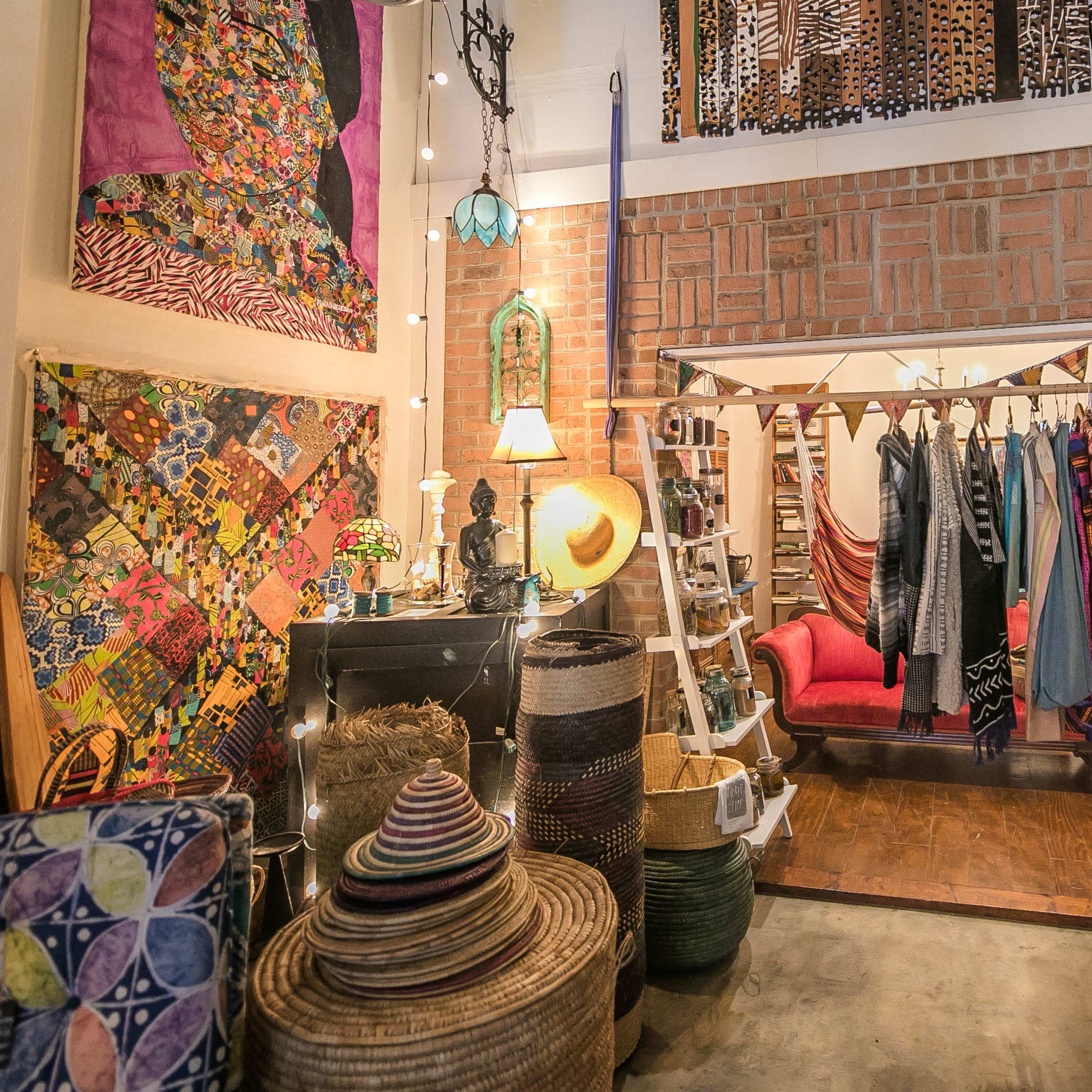 A cozy boutique displaying colorful textiles and clothing, with a brick wall backdrop and various woven baskets arranged around the room.