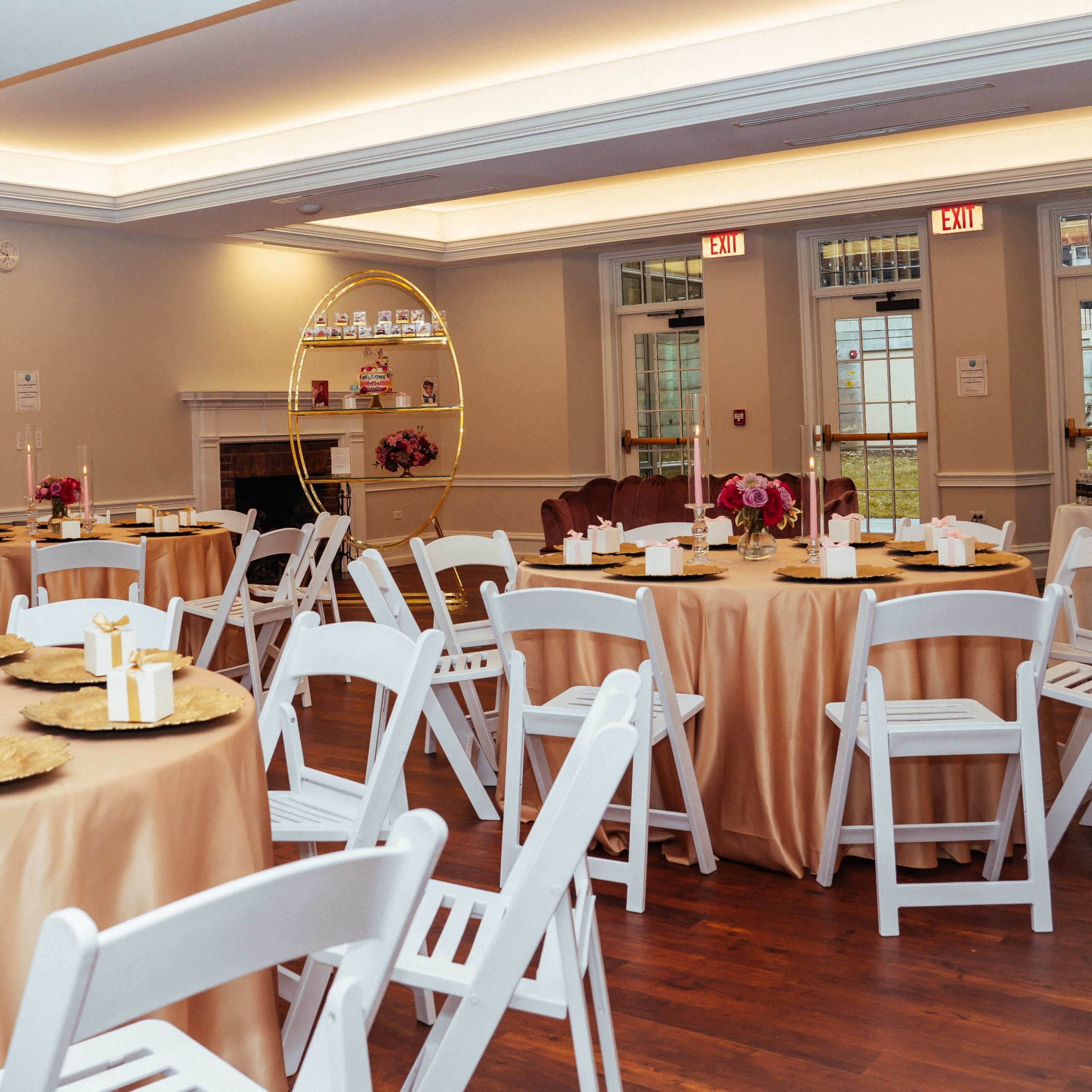 A banquet room set with round tables covered in gold and beige linens, each adorned with floral centerpieces and white chairs arranged around them.